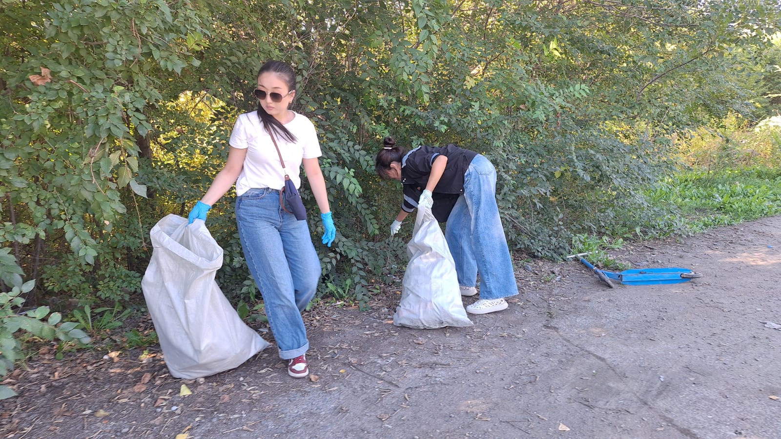 Екі қыз жол жиегіндегі қоқысты үлкен пакеттерге жинап жатыр.