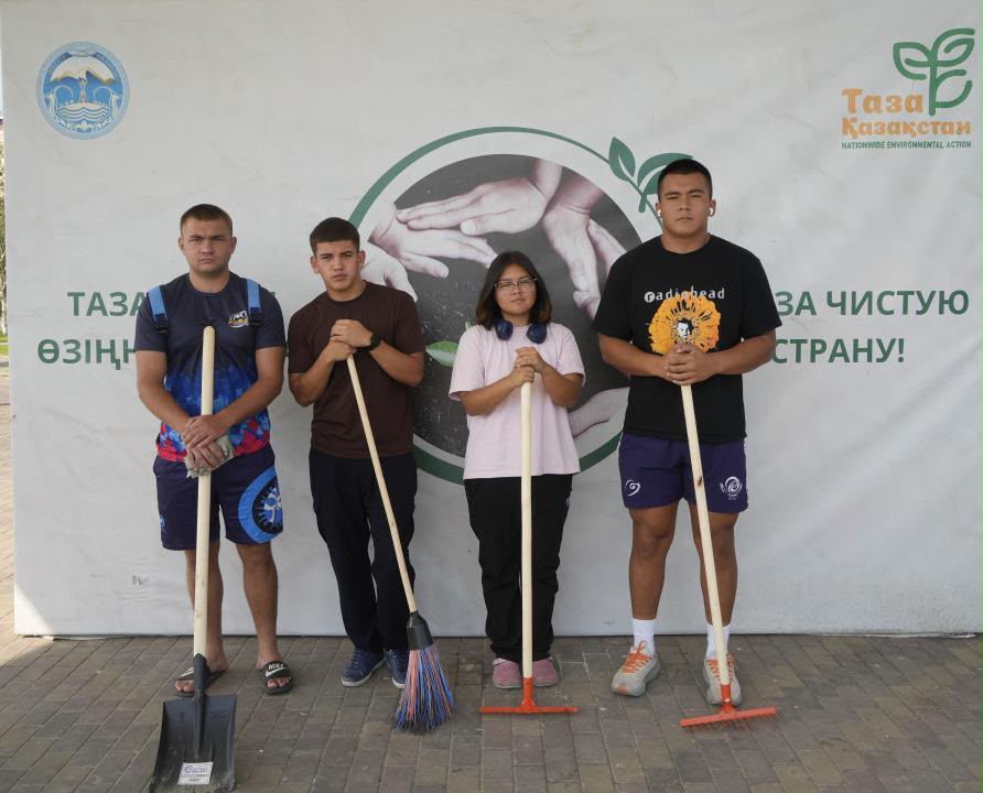 «Таза Қазақстан» акциясына қатысушы төрт жасөспірім қолындағы тазалау құралдарымен баннер алдында тұр.