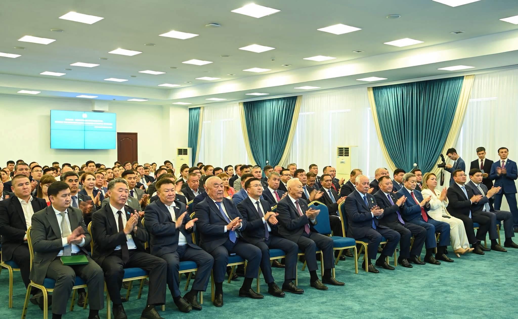 Конференция залында жиналған, қол шапалақтап отырған костюм киген ерлер мен әйелдер тобы.