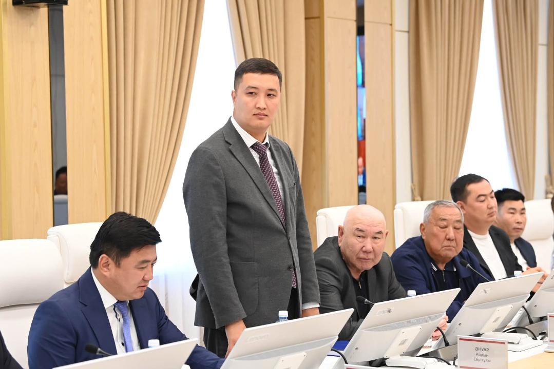 Жиналыста тұрған костюм киген ер адам және үстел басында отырған әріптестері.