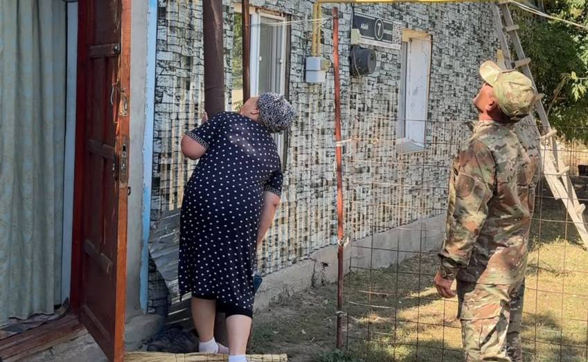 Үйдің жанында тұрған әскери киімдегі ер адам және нүктелі көйлек киген әйел.