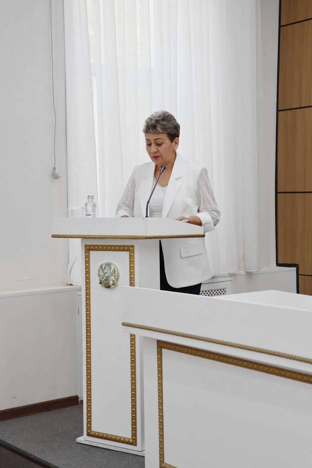 Ақ костюм киген әйел адам трибунада тұрып сөйлеп жатыр.