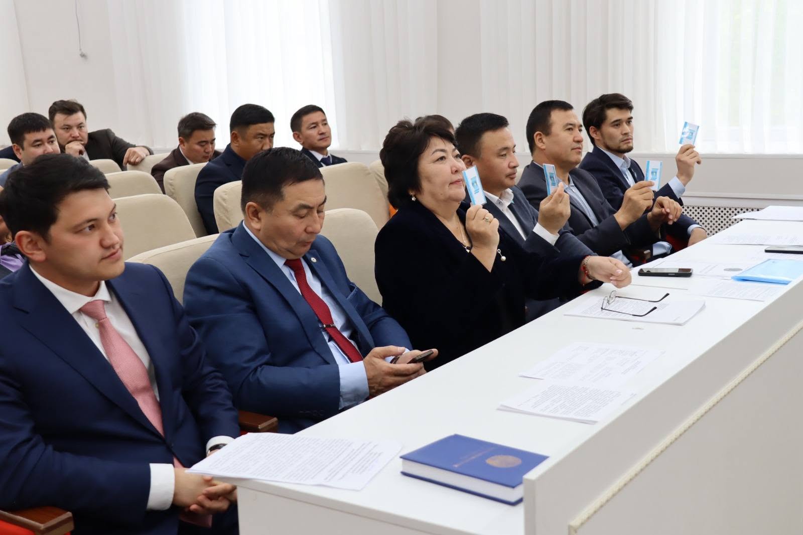 Костюм киген адамдар жиналыста отырып, қолдарымен дауыс беру карталарын көтеріп тұр.