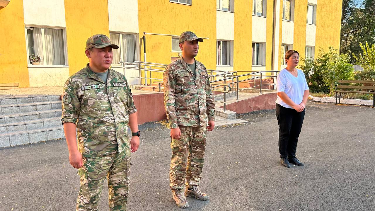 Сары ғимараттың алдында тұрған екі әскери және бір әйел.