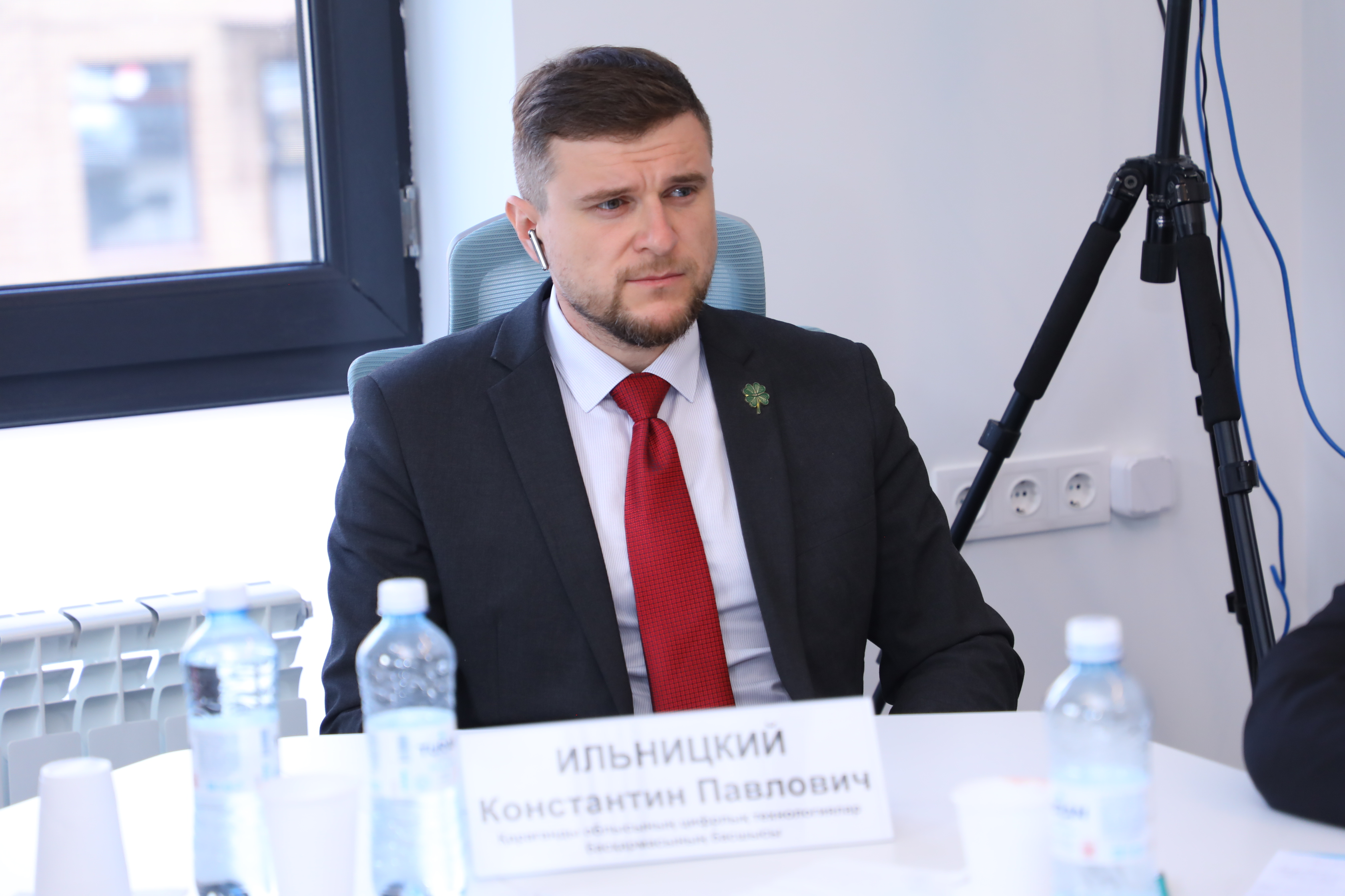 Қара костюм мен қызыл галстук киген, алдында есімі жазылған тақтасы бар ер адам.