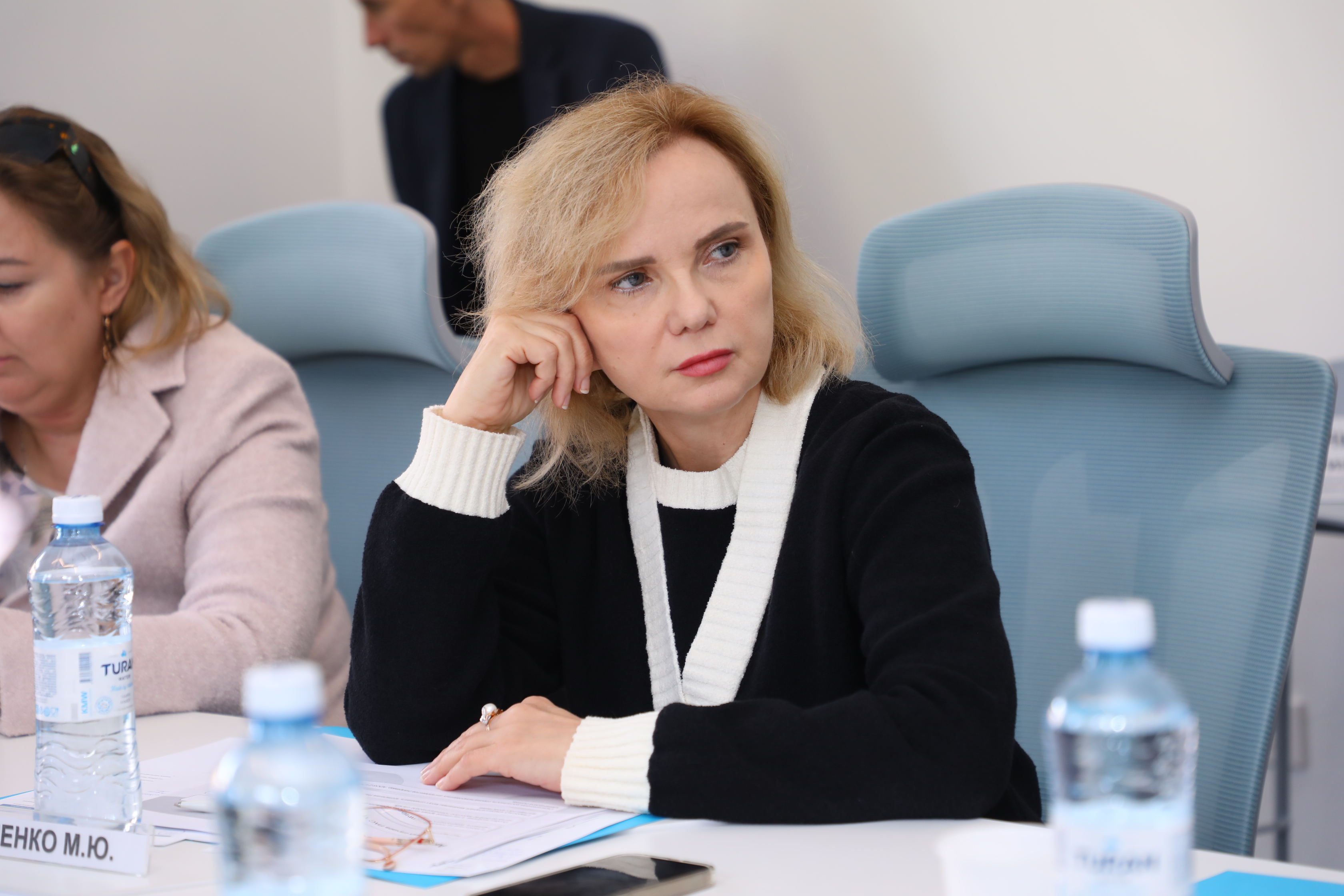 Көк орындықта отырған, ойлы көңіл-күйдегі әйел. Алдында су бөтелкелері мен құжаттар тұр.