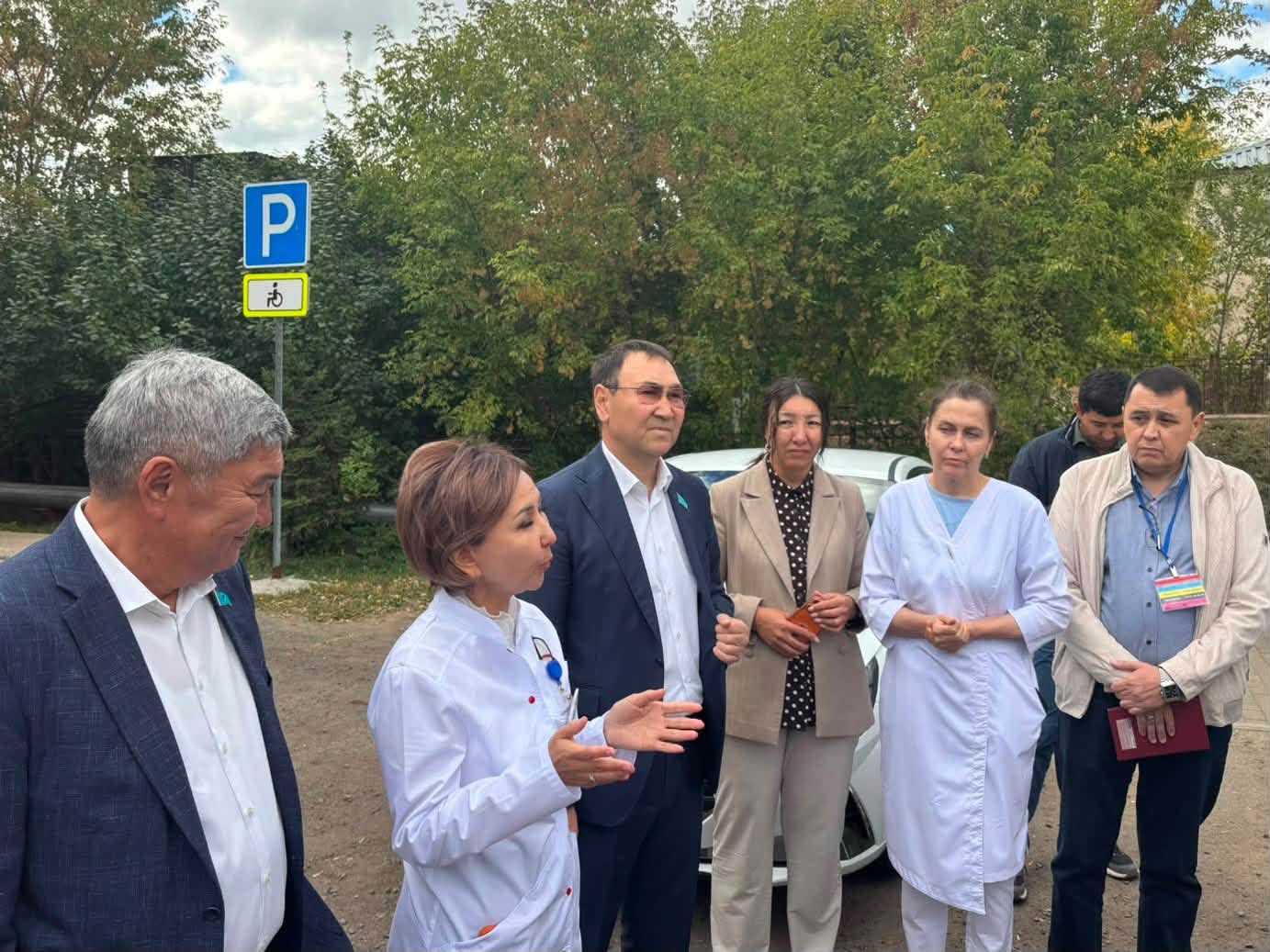 Сыртқы жерде жиналған ресми киімдегі және медициналық халат киген адамдар тобы.