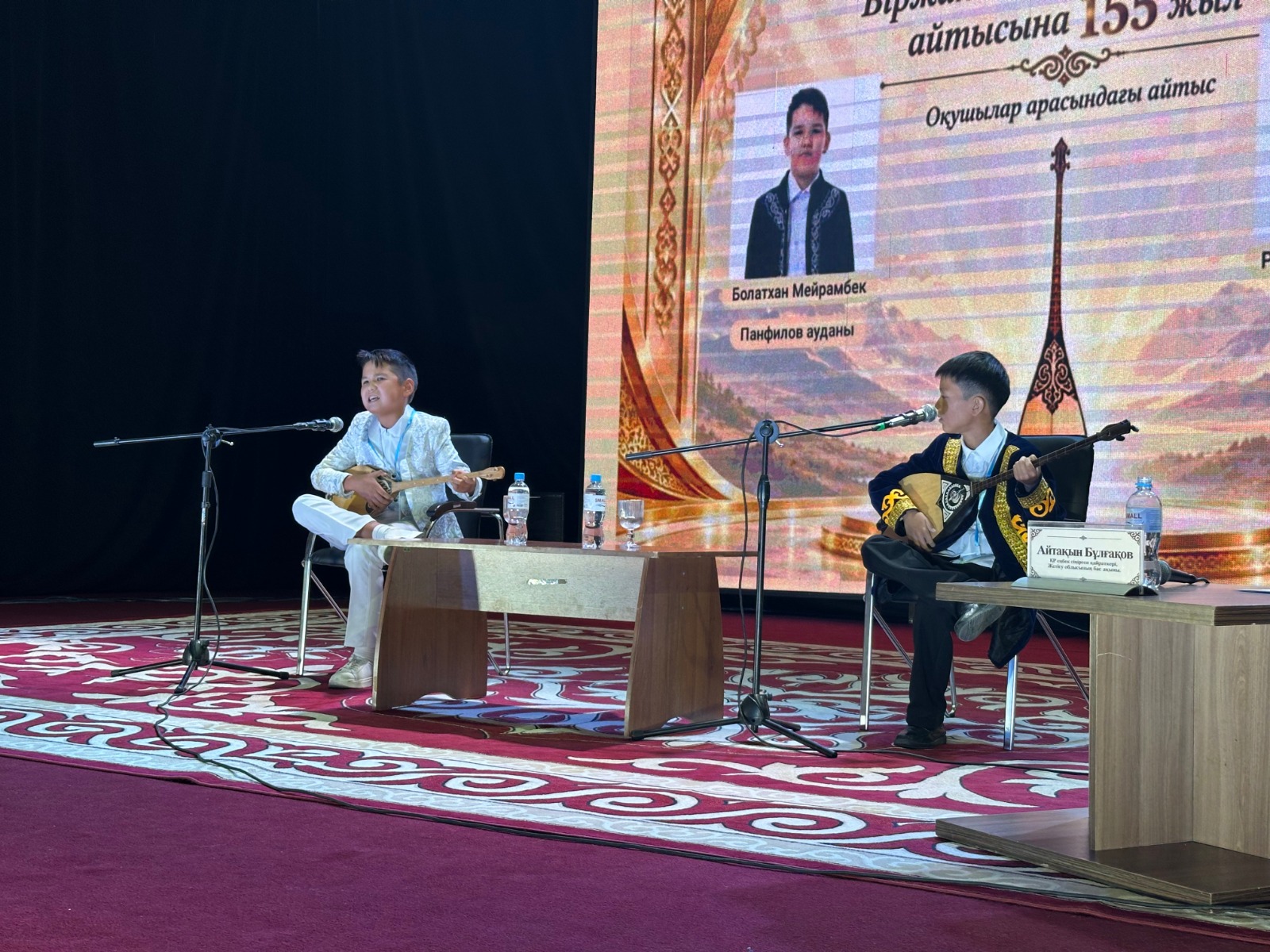 Сахнада домбыра тартып, айтысып жатқан екі бала.