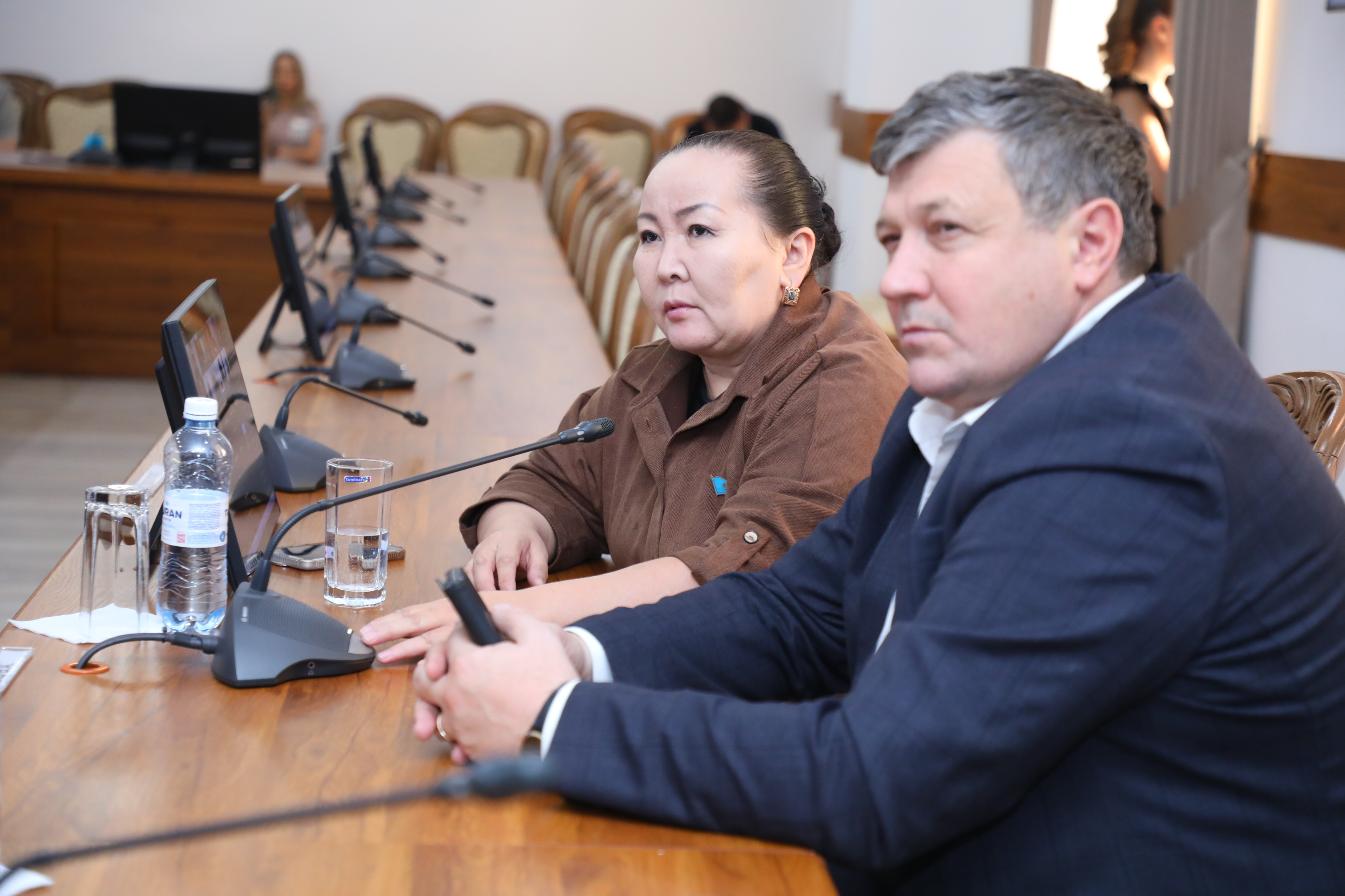 Жиналыс үстіндегі екі адам: костюм киген ер адам және қоңыр киімдегі әйел. Үстелде микрофондар мен су мен стақандар тұр.