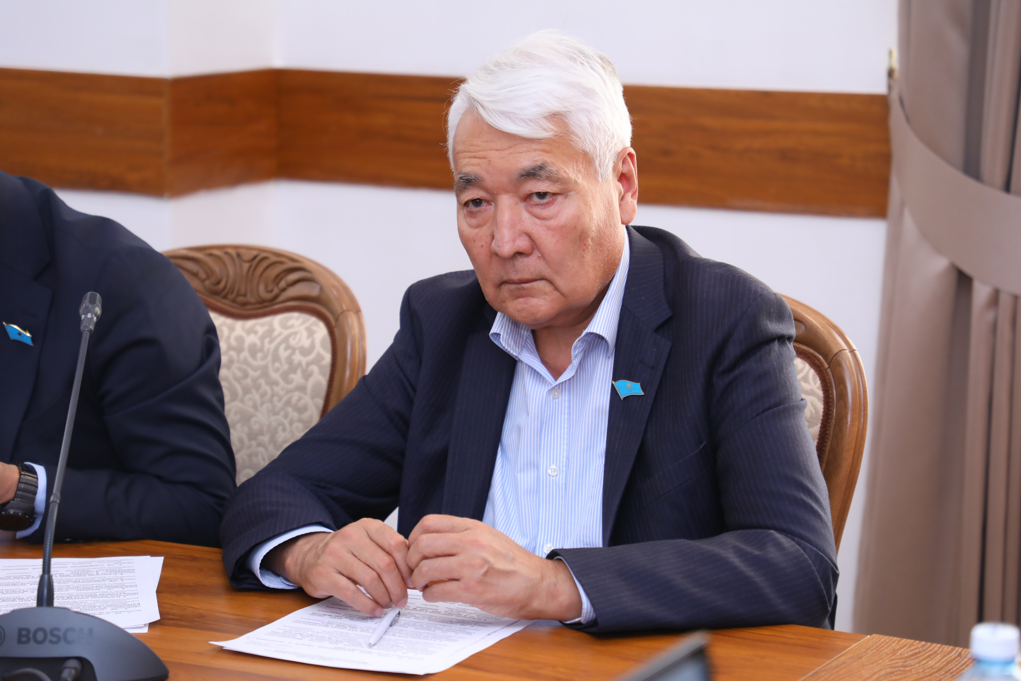 Көк костюм киген, ақ шашты ер адам үстел басында құжаттармен жұмыс істеп отыр.