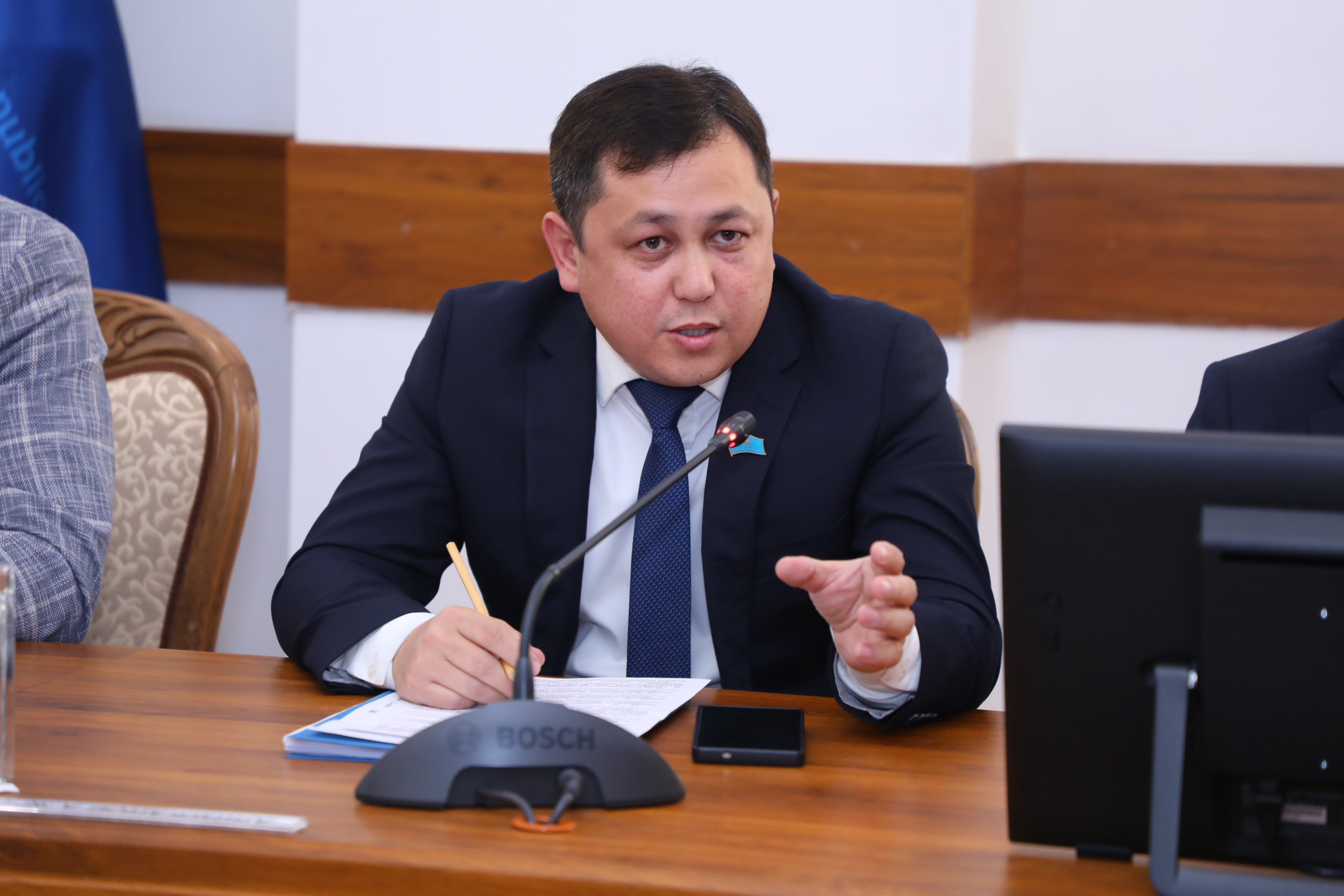 Көк костюм киген ер адам жиналыста сөйлеп тұр.