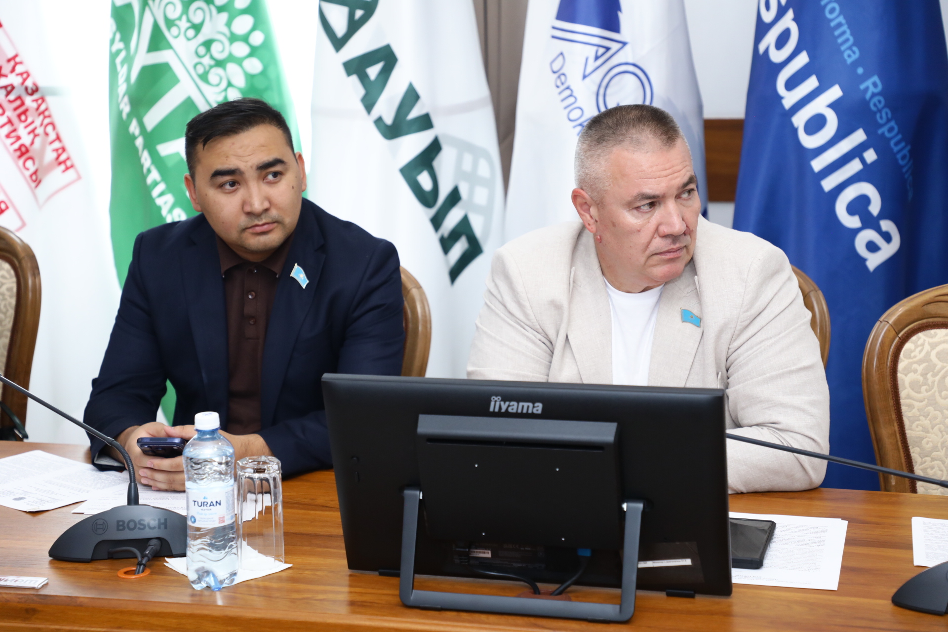 Жиналыста отырған екі депутат және артқы жақтағы әртүрлі партиялардың тулары.