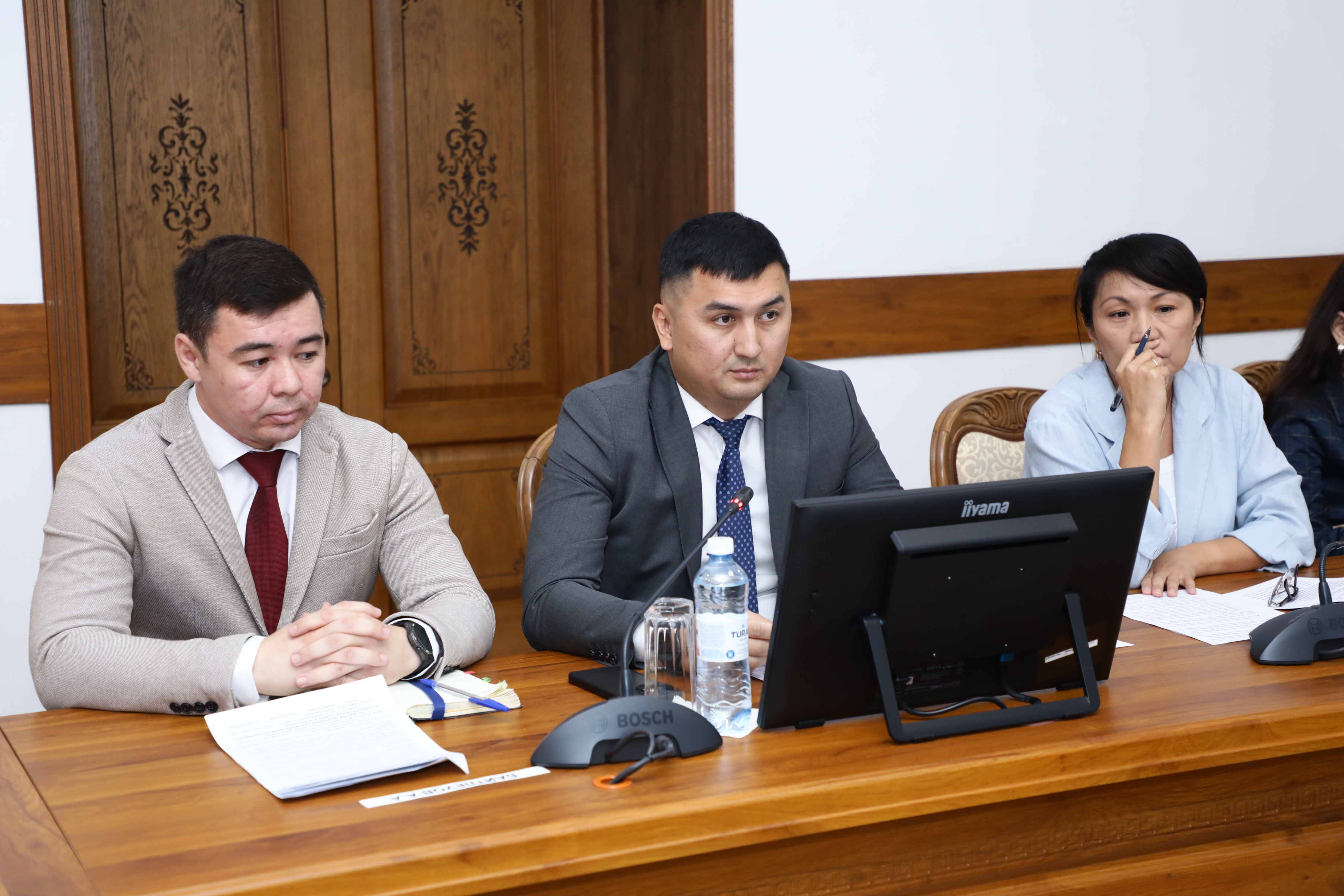 Жылынау бөлмеде үстел басында отырған үш ресми киінген адам.