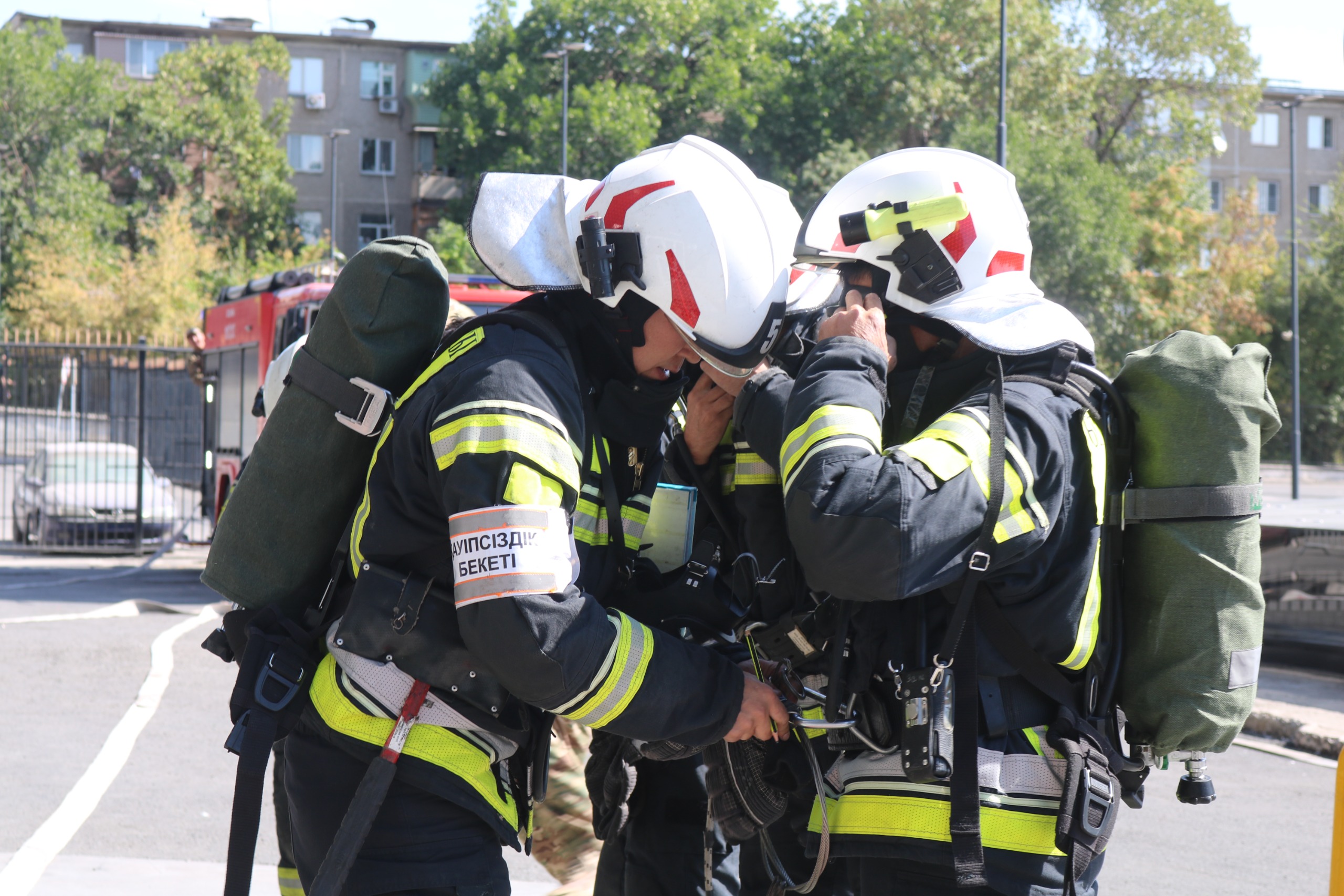 Двое пожарных в полной экипировке и белых касках проверяют снаряжение друг друга на фоне пожарной машины.