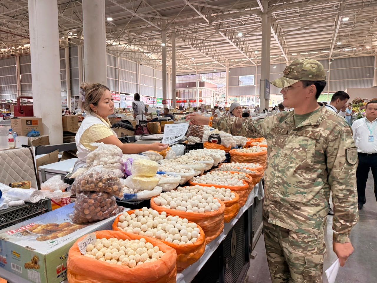Военный в камуфляже выбирает курт и другие молочные продукты на рынке, общаясь с продавщицей.