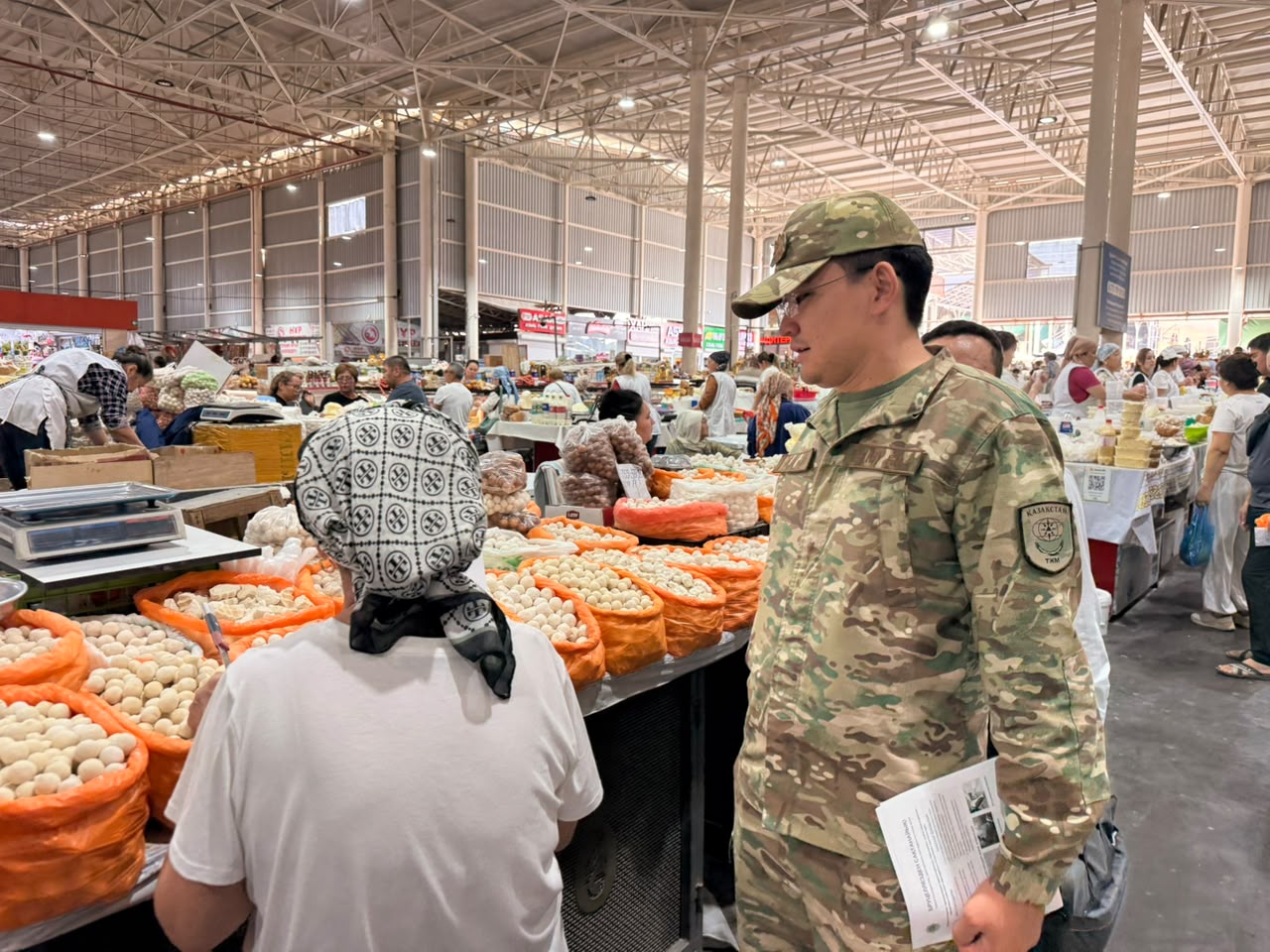 Военный в камуфляже общается с женщиной-продавцом на рынке с мешками белых продуктов.