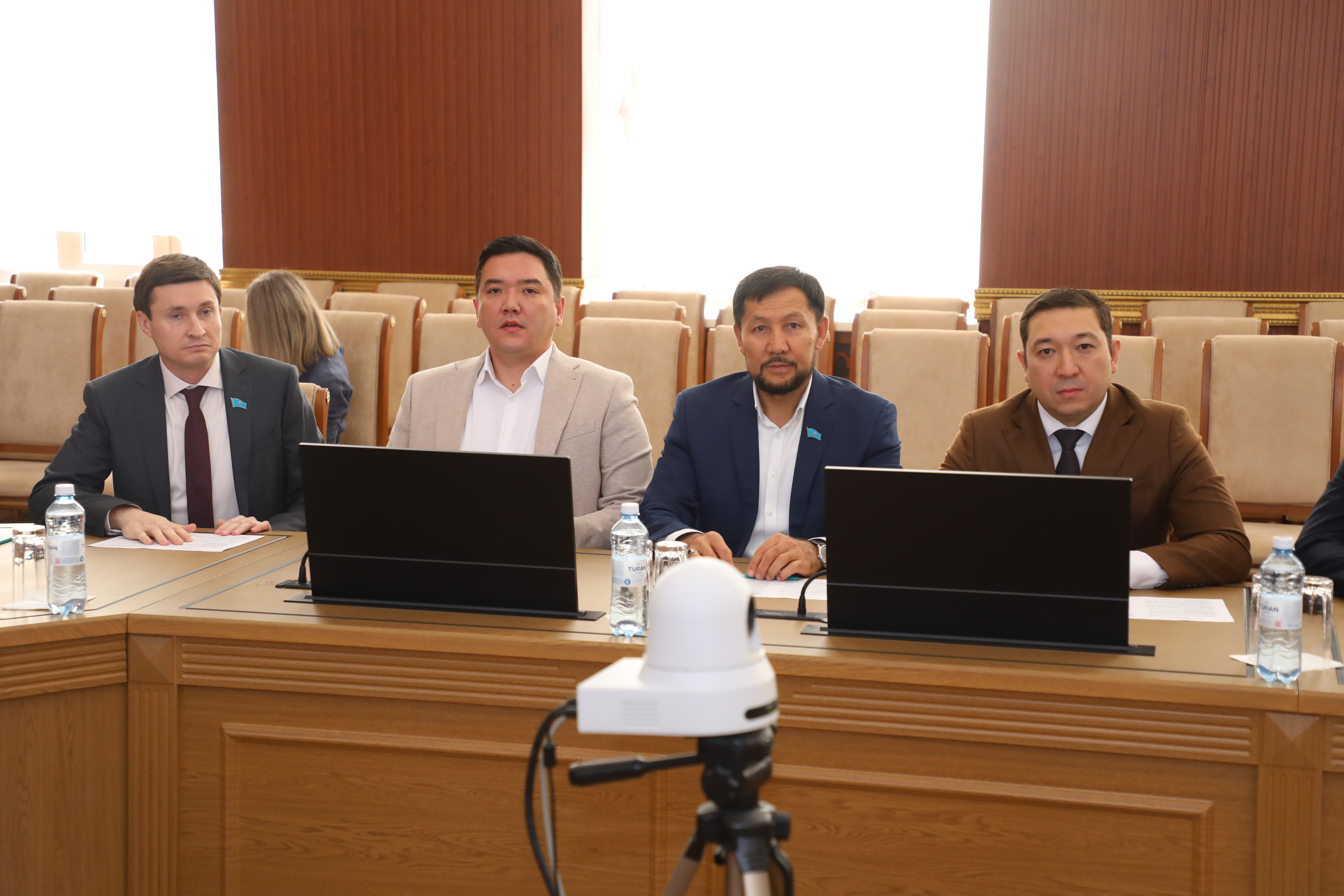 Кеңес залында ноутбуктер мен микрофонның алдында отырған төрт ер адам.