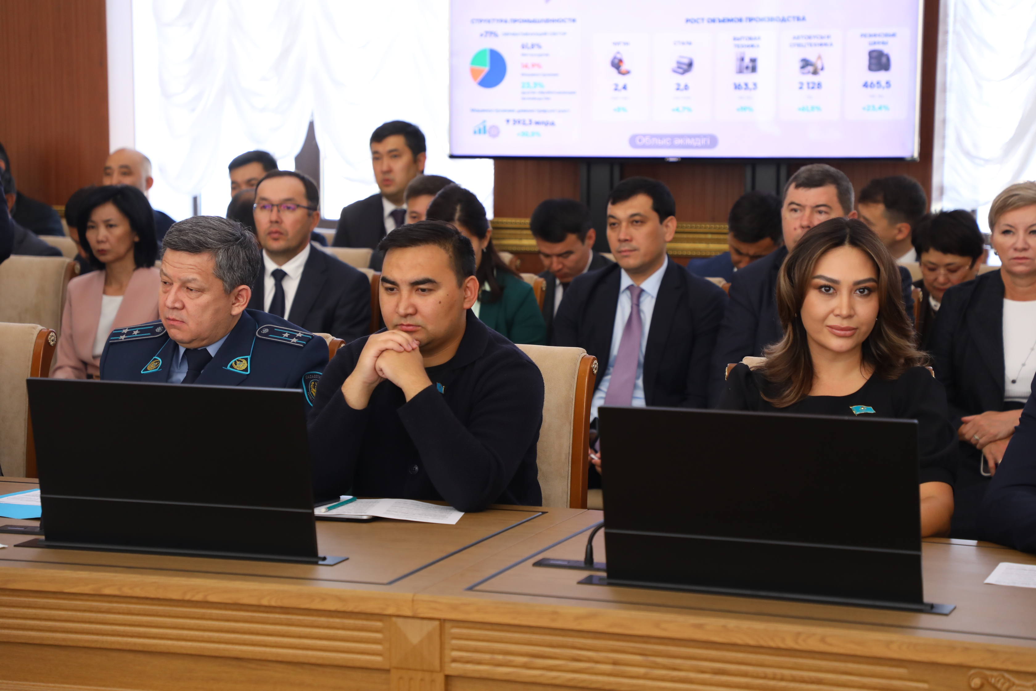 Конференцияда ноутбуктердің алдында отырған ресми киінген адамдар тобы.