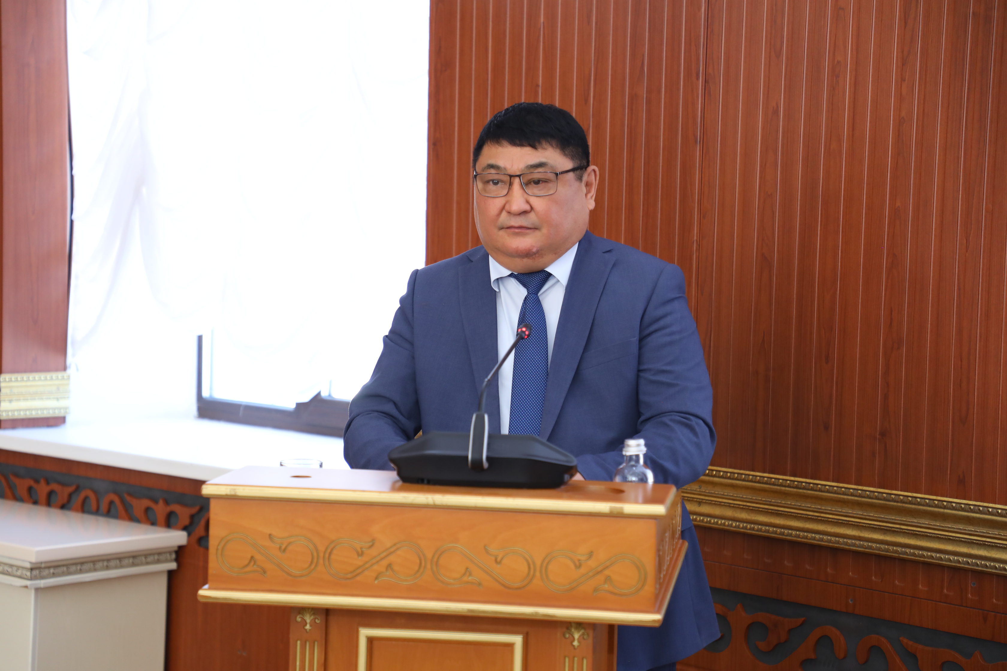 Көзілдірік пен көк костюм киген ер адам трибунада сөйлеп тұр.
