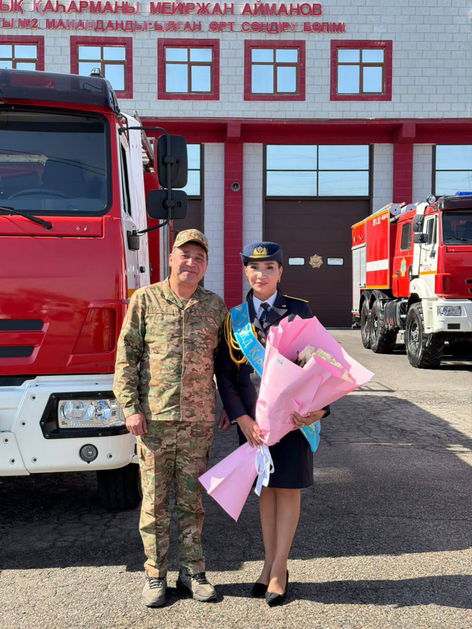 Өрт сөндіру бөлігінің алдында әскери формадағы ер адам мен гүл ұстаған формадағы әйелдің суреті.