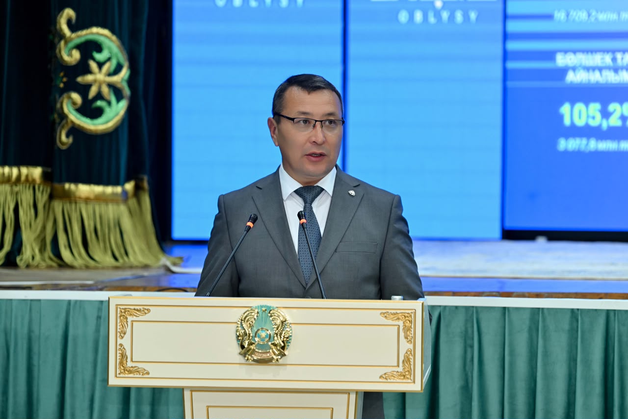 Көзәйнектің алдында тұрған, костюм киген ер адамның сөйлеп жатқан сәті.