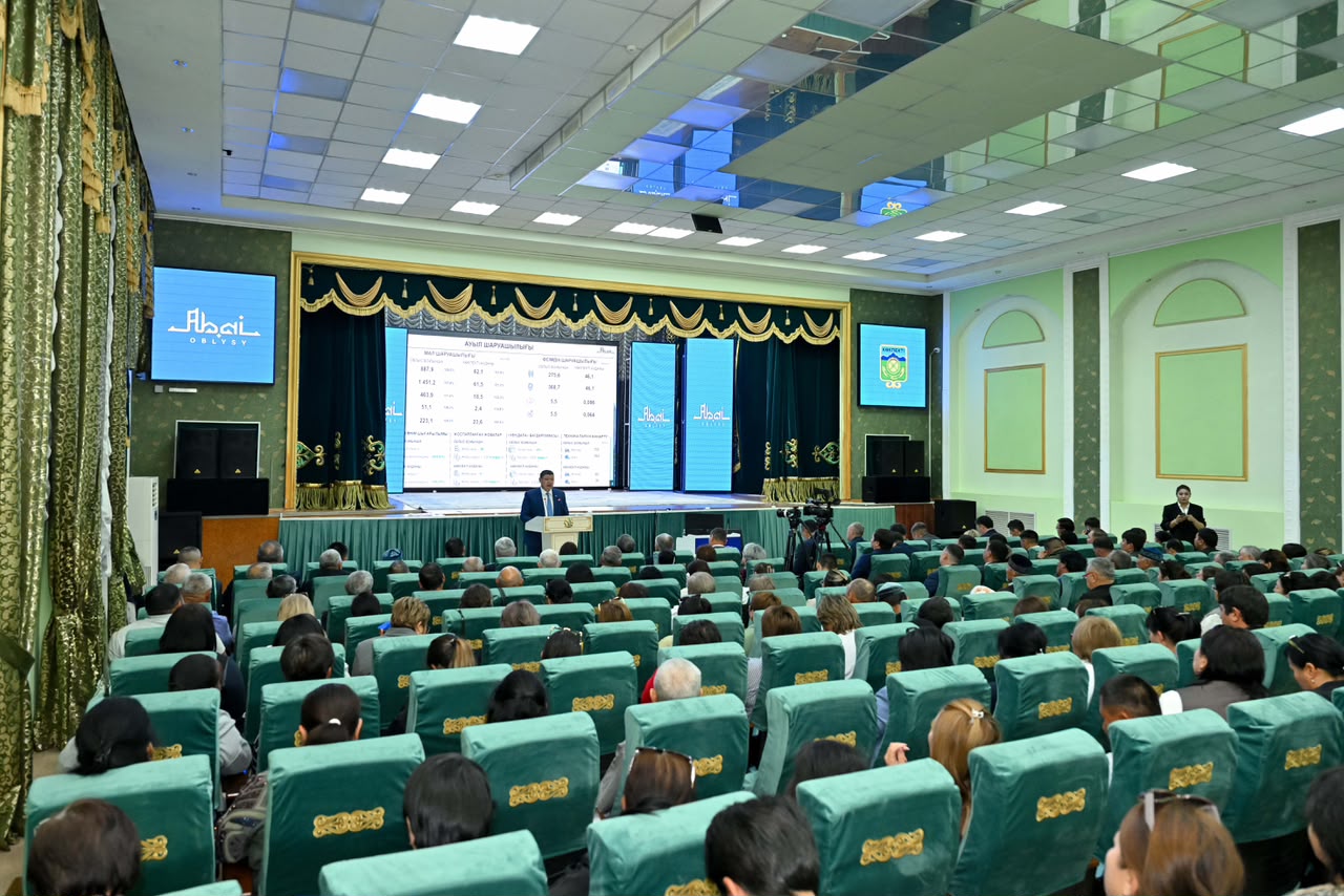 Конференция залындағы баяндамашы мен тыңдаушылар. Экранда статистикалық мәліметтер көрсетілген.