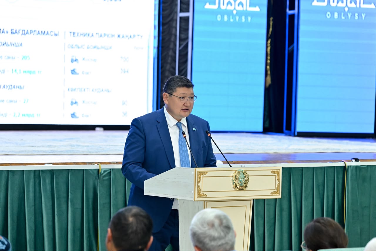 Көк костюм киген ер адам трибунада сөйлеп тұр.