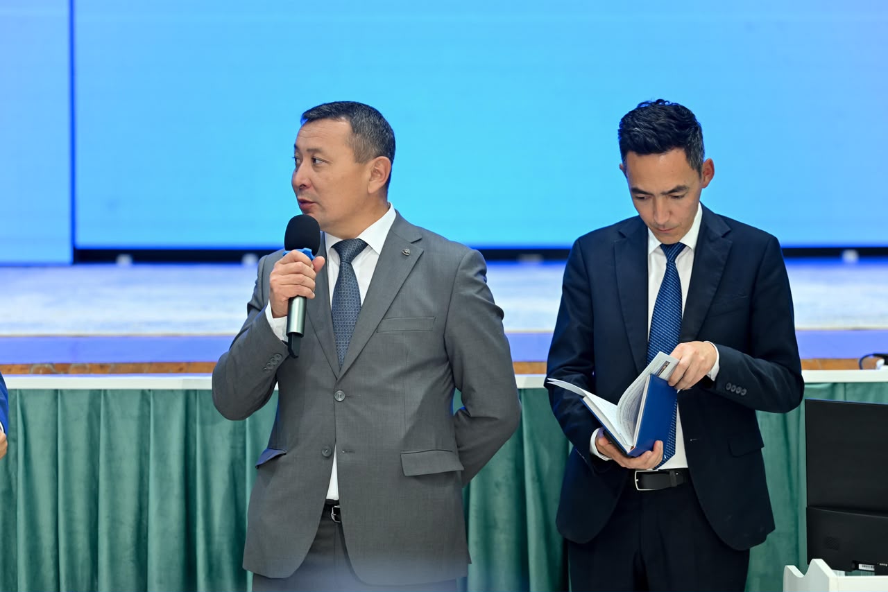 Микрофонмен сөйлеп тұрған костюмді ер адам және қасында кітапша қарап тұрған әріптесі.