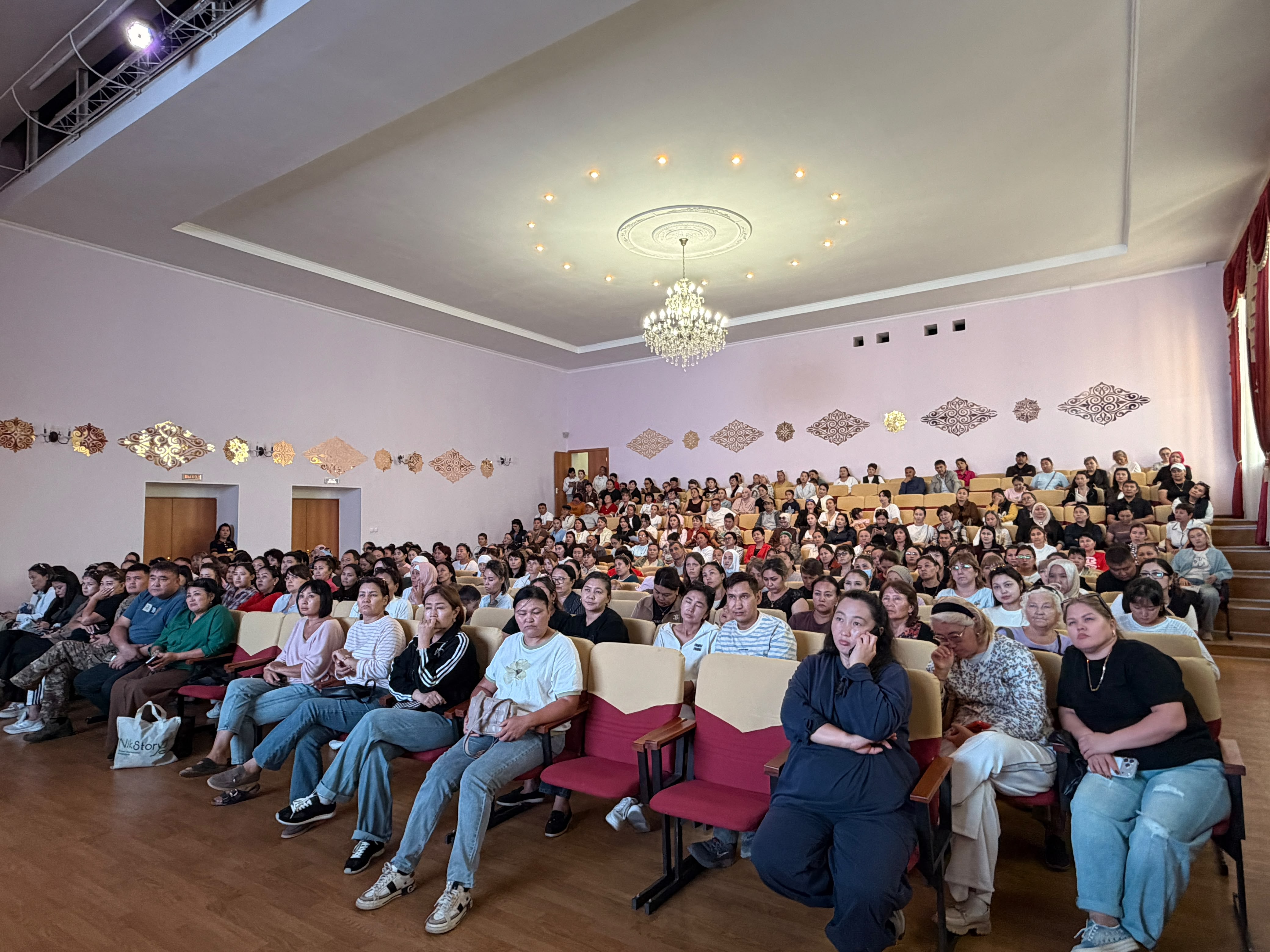 Зрители в заполненном зале с люстрой и декоративными панно на стенах.
