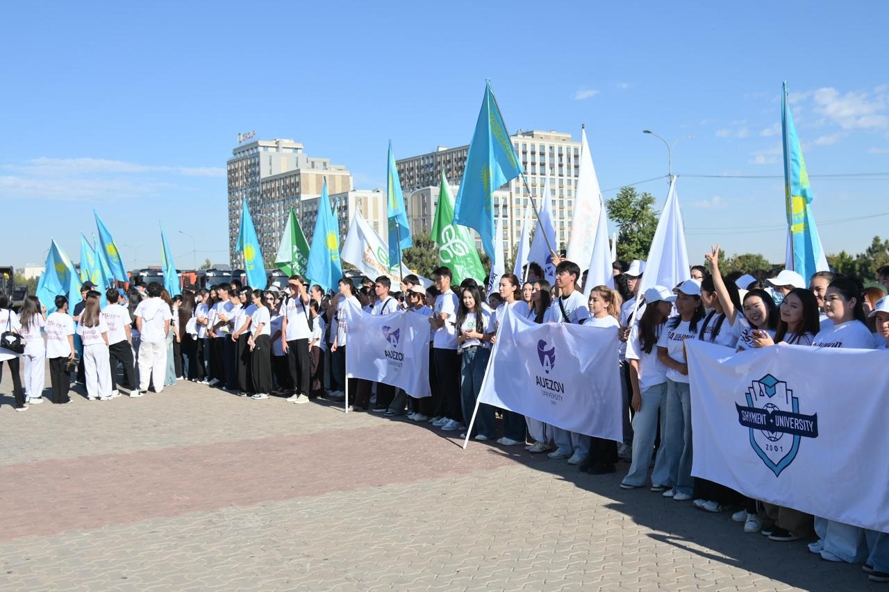 Ақ киімді студенттердің Қазақстан туларымен және университет баннерлерімен жиналған топтық суреті.
