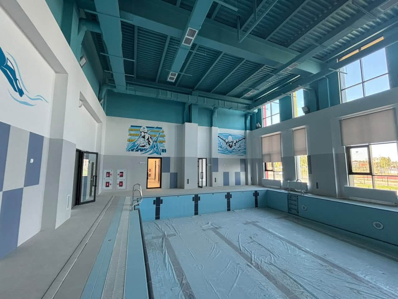 Көк түсті интерьері бар, қабырғаларында жүзушілердің суреттері бейнеленген жабық бассейн.