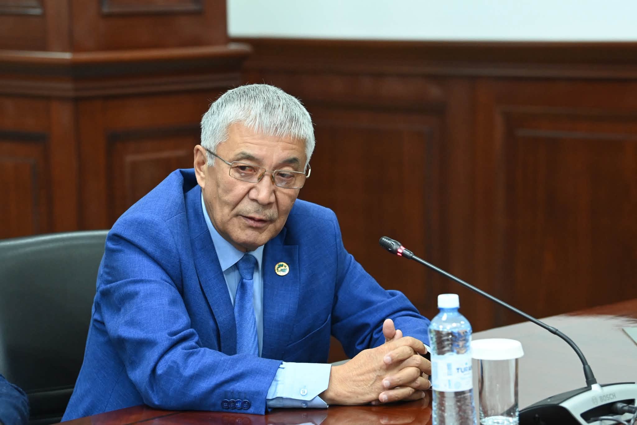 Көк костюм киген, көзілдірікті ер адам жиналыста сөйлеп тұр.