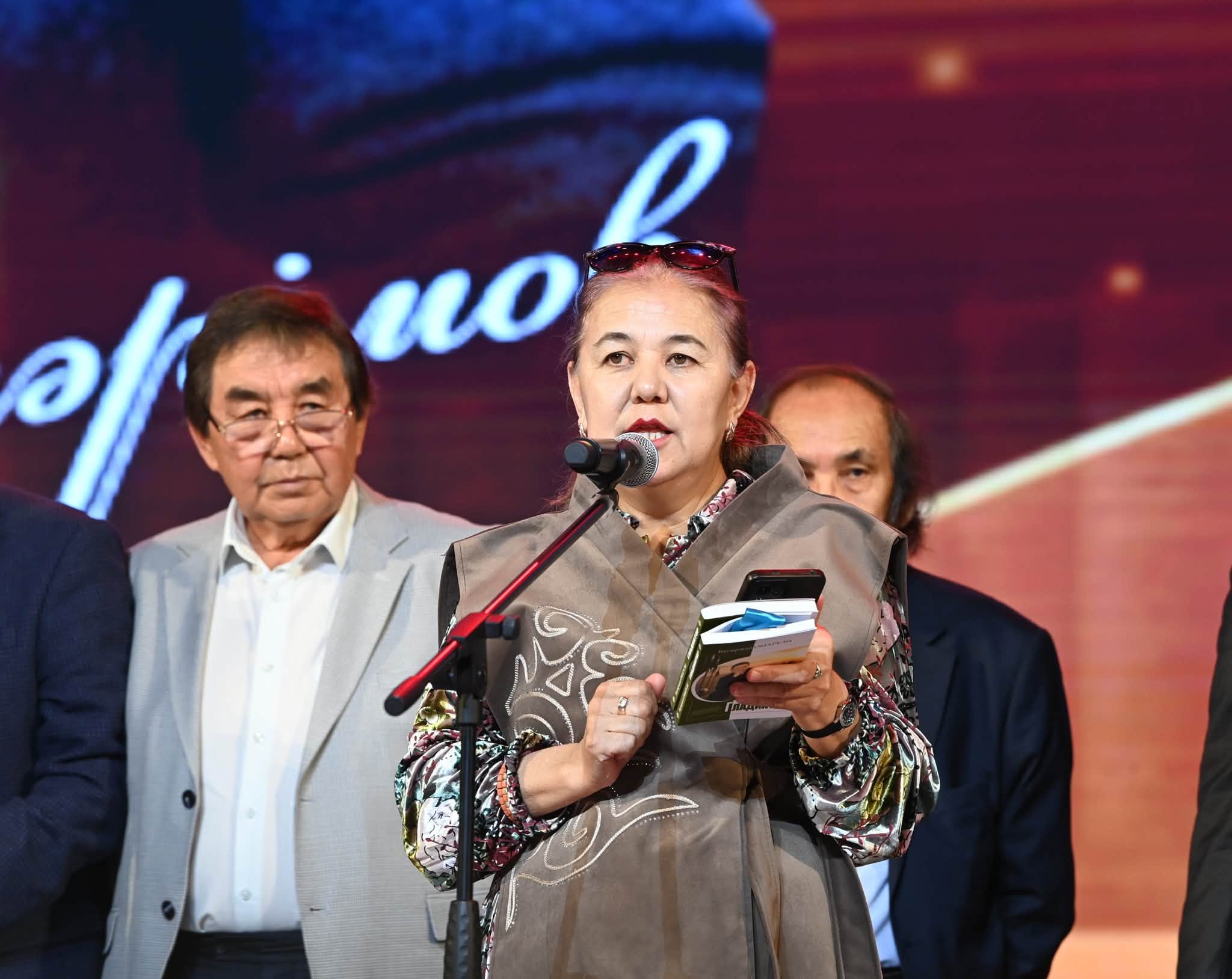 Микрофонда сөйлеп тұрған, қолында кітап ұстаған әйел.