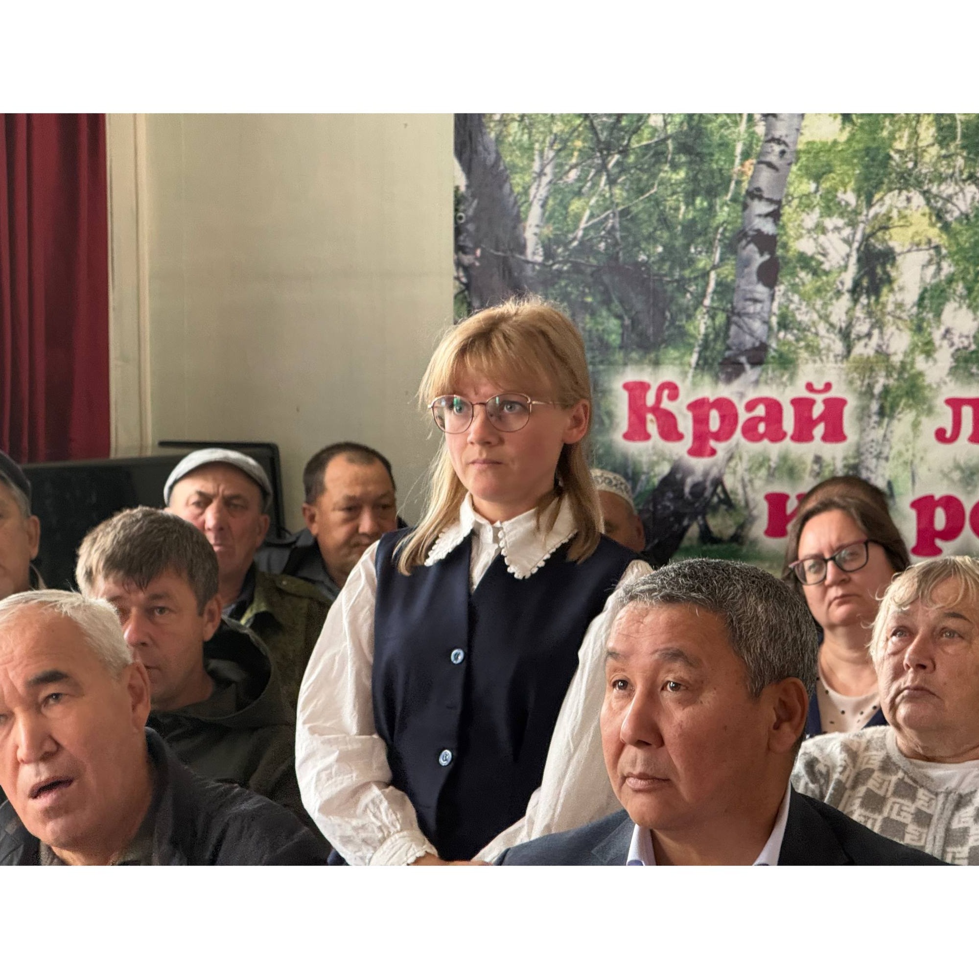 Женщина в очках и синем жилете сидит в группе людей