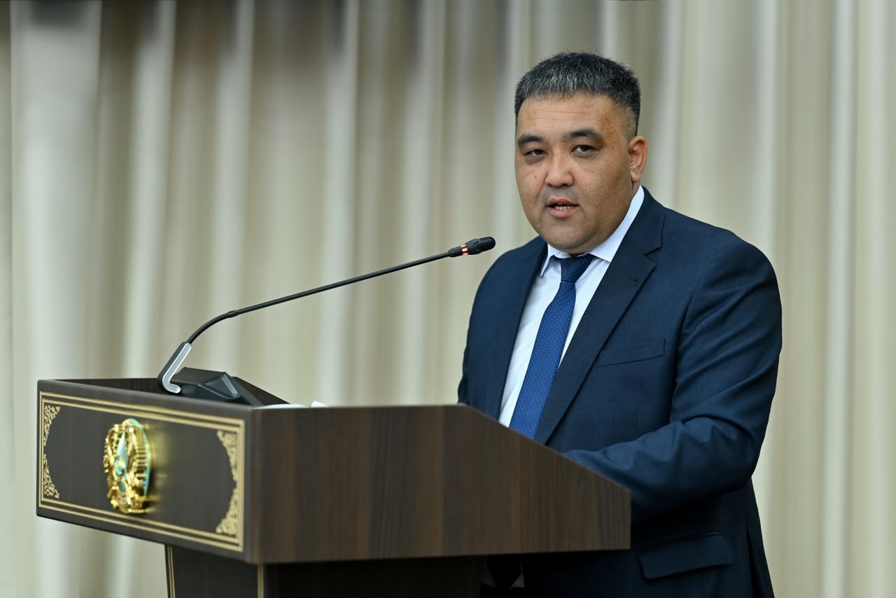 Көк костюм киген ер адам трибунада сөйлеп тұр.