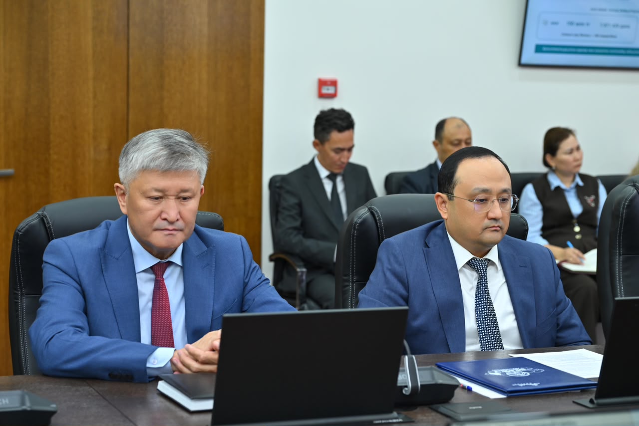 Көк костюм киген екі ер адам жиналыста ноутбук пен құжаттарға қарап отыр.