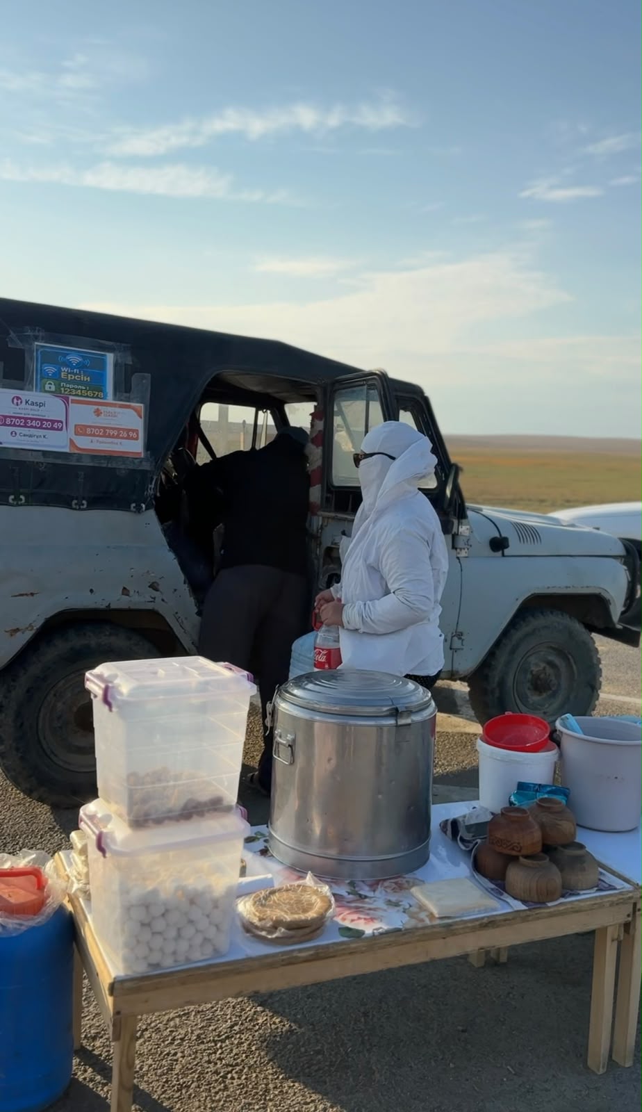 Жол жиегіндегі сауда нүктесі: үстелдегі тағамдар мен ақ киінген сатушы, артқы жағында ақ түсті жол көлігі.