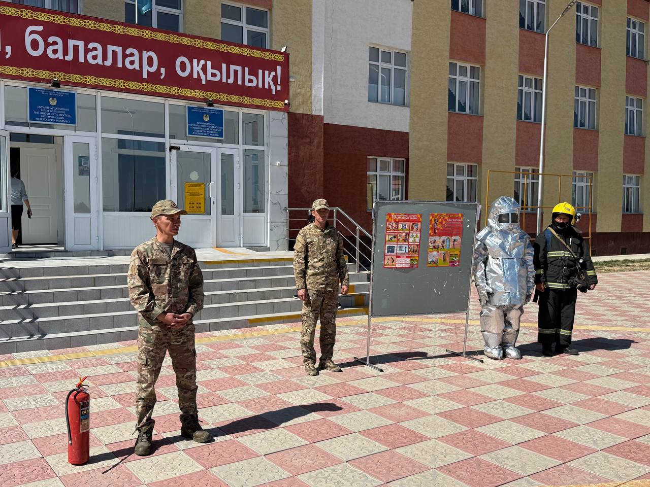 Мектеп алдында тұрған әскери қызметшілер мен өрт сөндірушілер. Жанында өрт сөндіргіш пен ақпараттық стенд тұр.