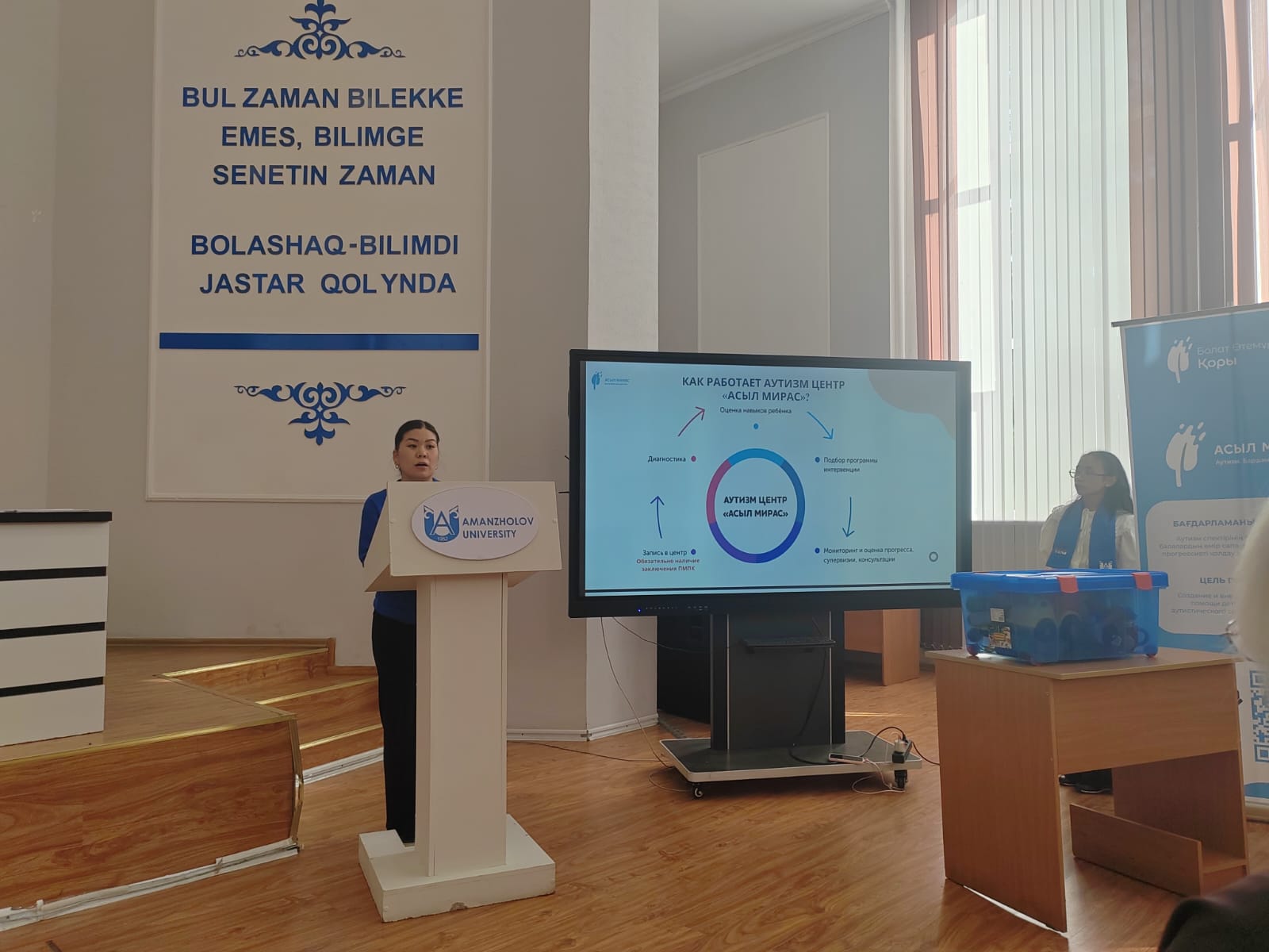 Amanzholov университетіндегі презентация: спикер экрандағы аутизм орталығының жұмыс схемасын түсіндіріп тұр.