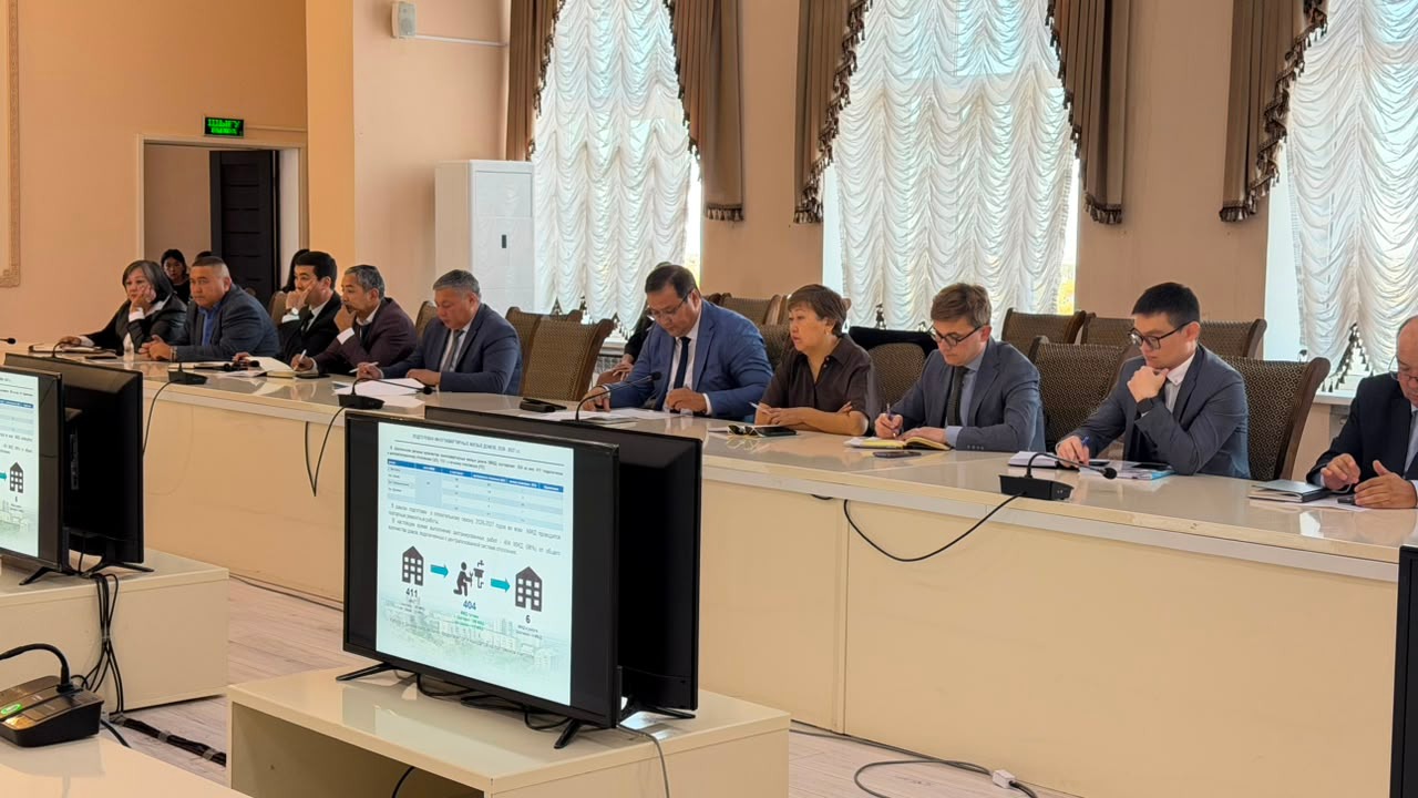 Іскерлік костюм киген адамдар тобы кеңес кезінде ұзын үстелде отыр, олардың алдында деректер көрсетілген мониторлар тұр.