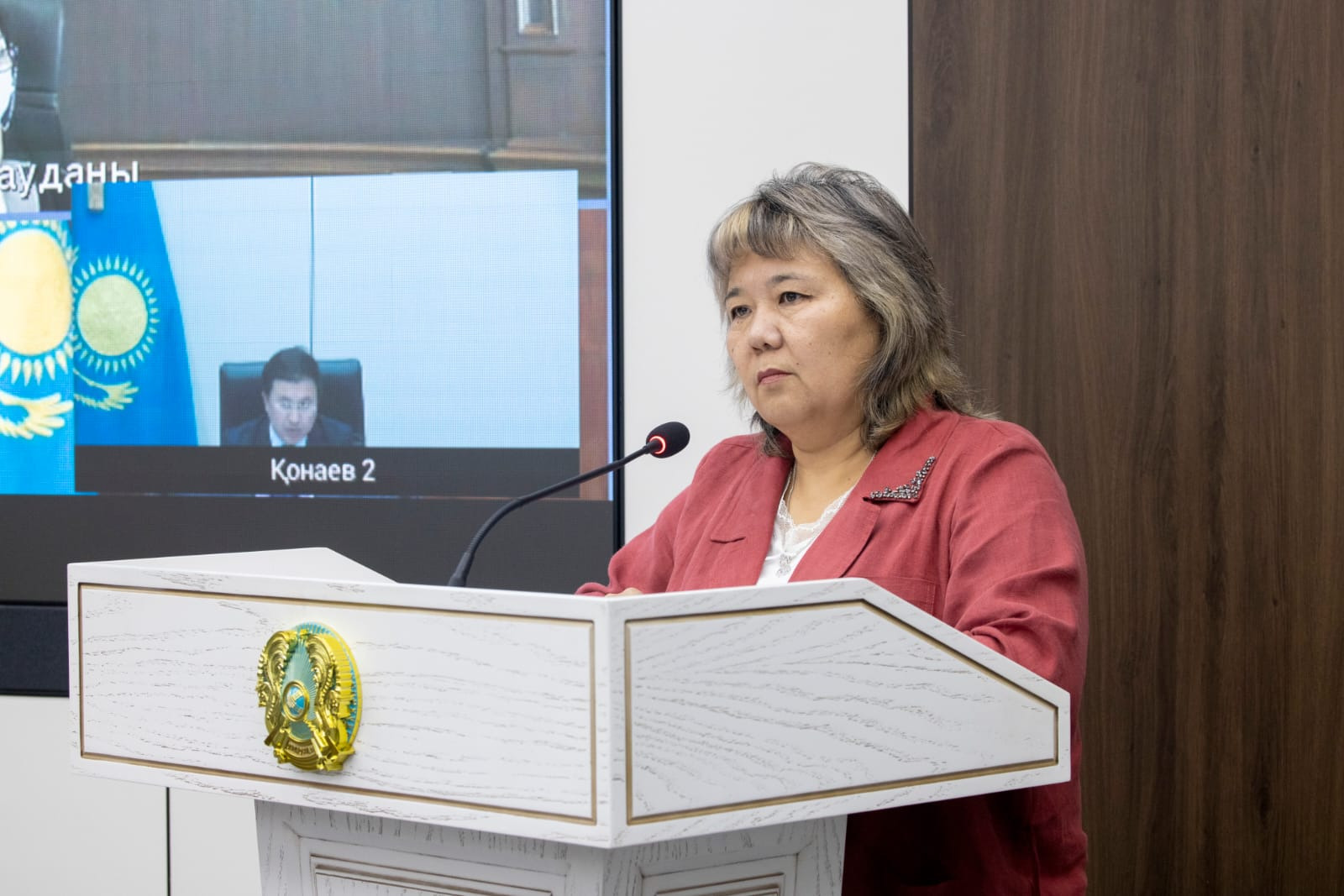 Трибунада сөйлеп тұрған әйел. Артында бейнебайланыс экраны мен Қазақстан Республикасының елтаңбасы бар.