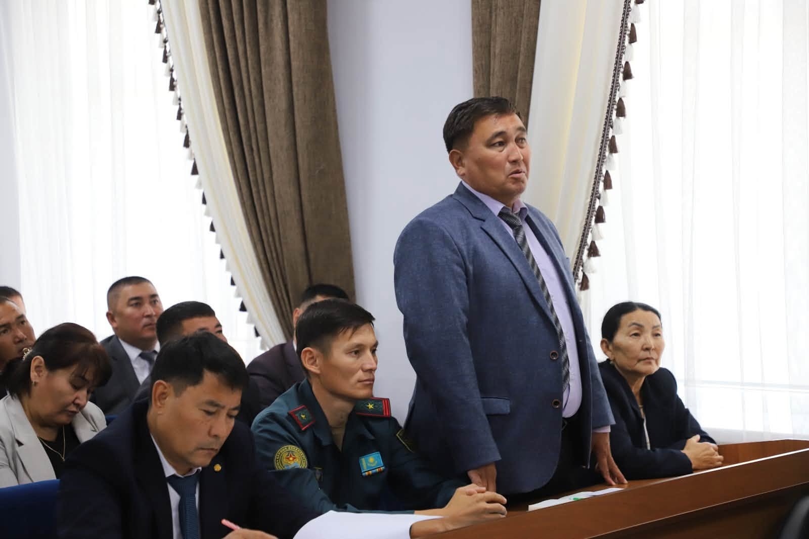 Группа мужчин в деловых костюмах на официальном мероприятии в конференц-зале. Один мужчина стоит и выступает перед сидящими.