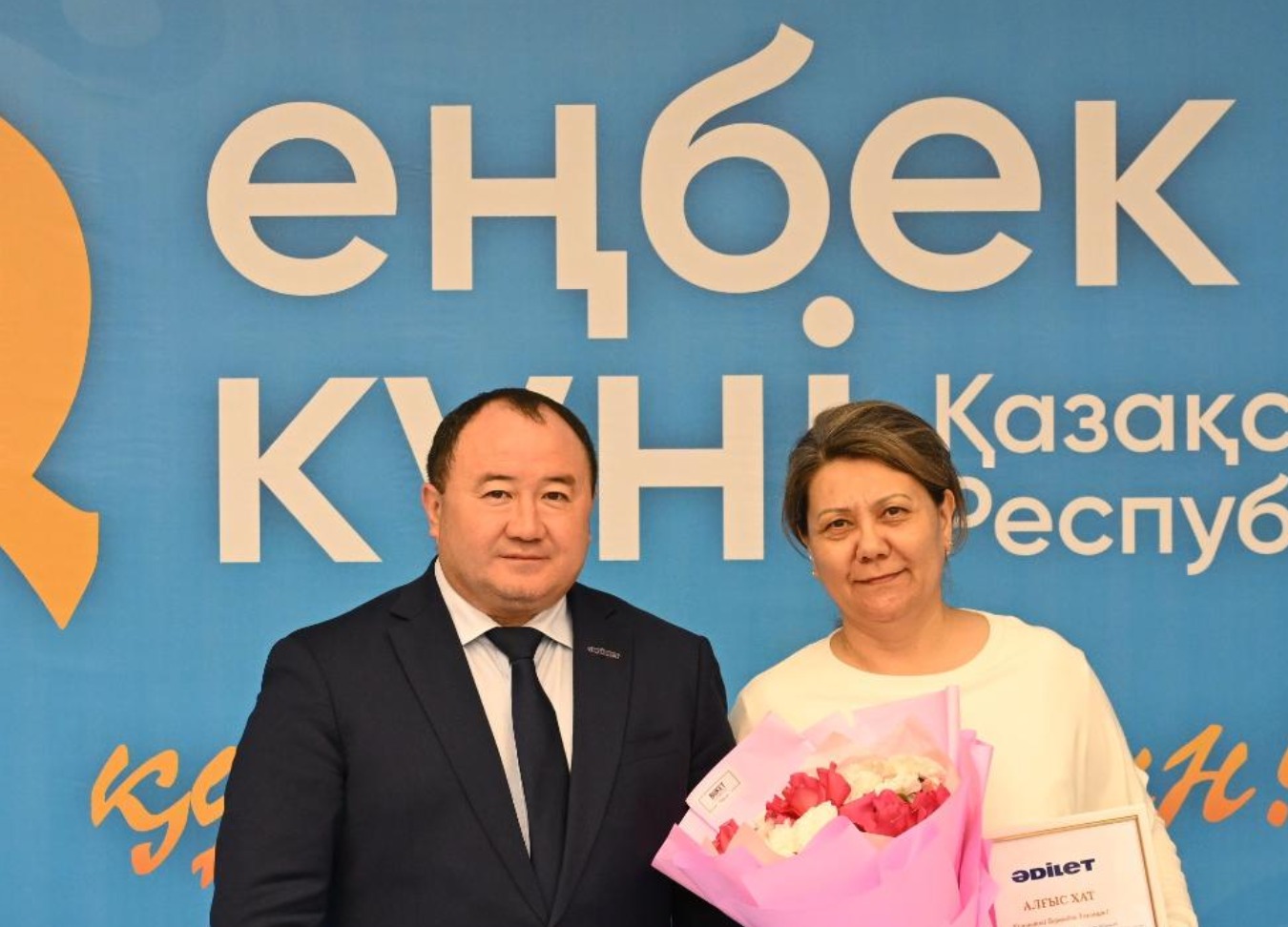 «Еңбек күні» баннерінің алдында тұрған, қолына гүл ұстаған әйел мен ер адам.