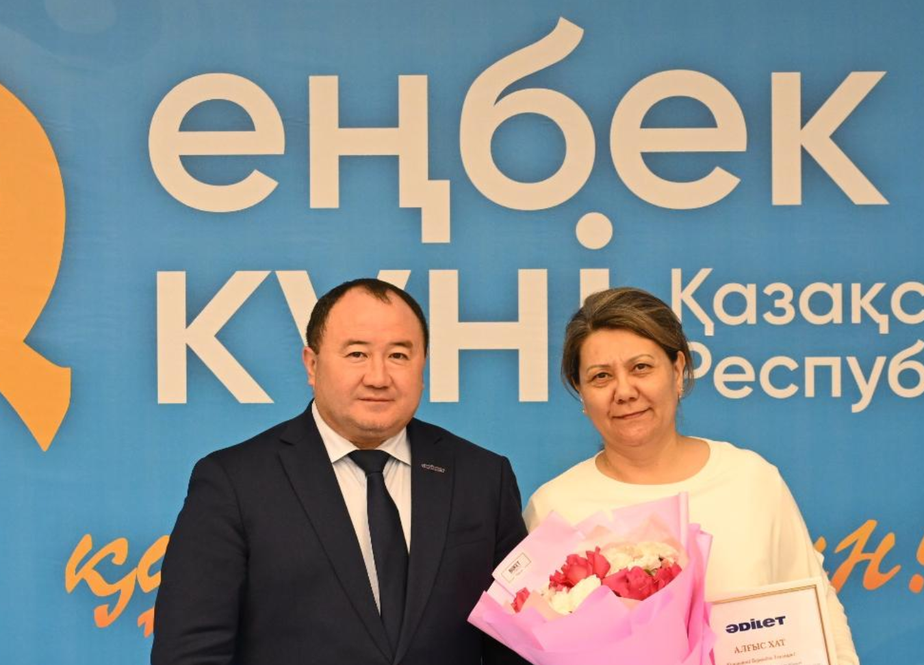 «Еңбек күні» баннерінің алдында тұрған, қолына гүл мен грамота ұстаған әйел мен костюм киген ер адам.