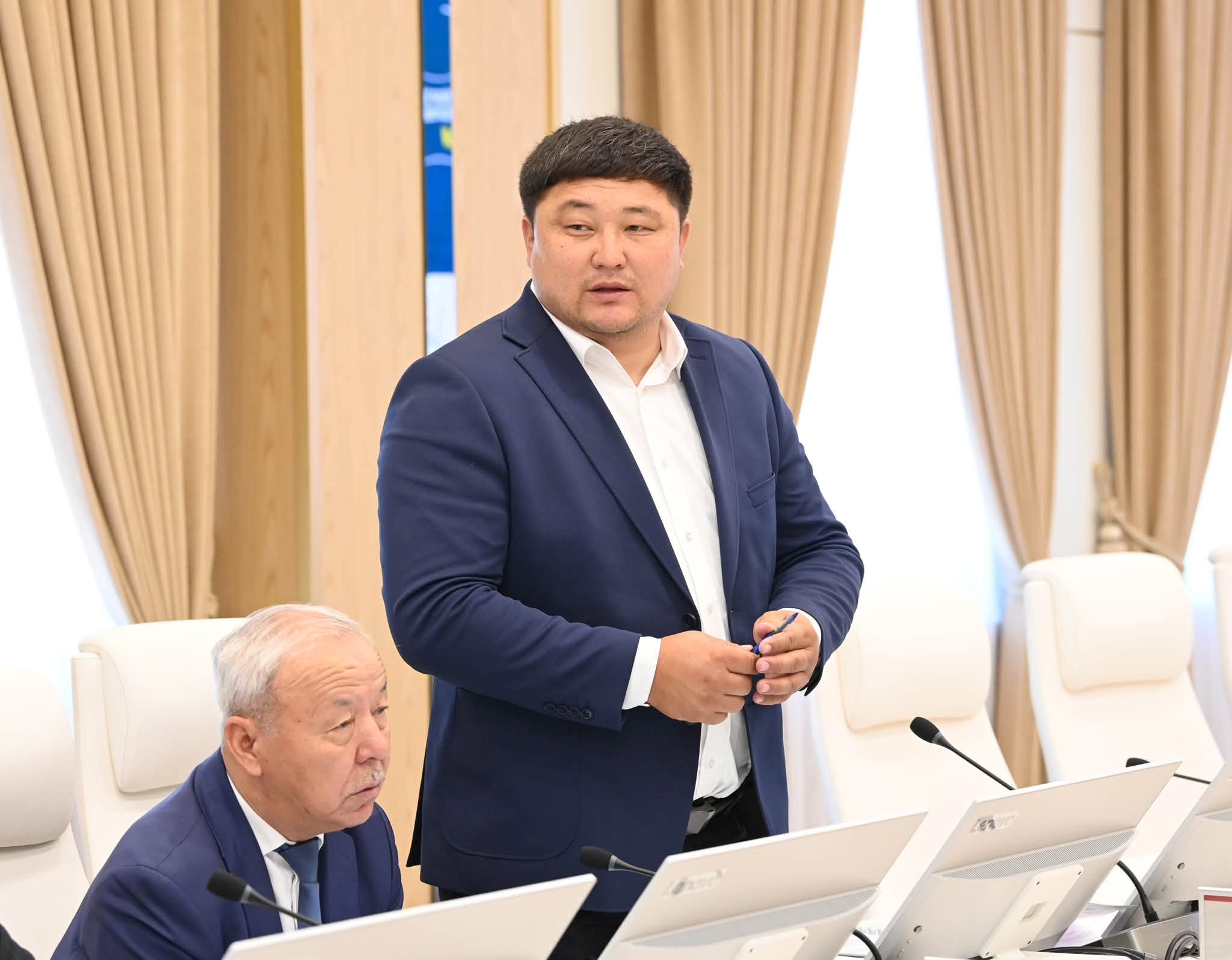 Көк костюм киген ер адам жиналыста сөйлеп тұр.