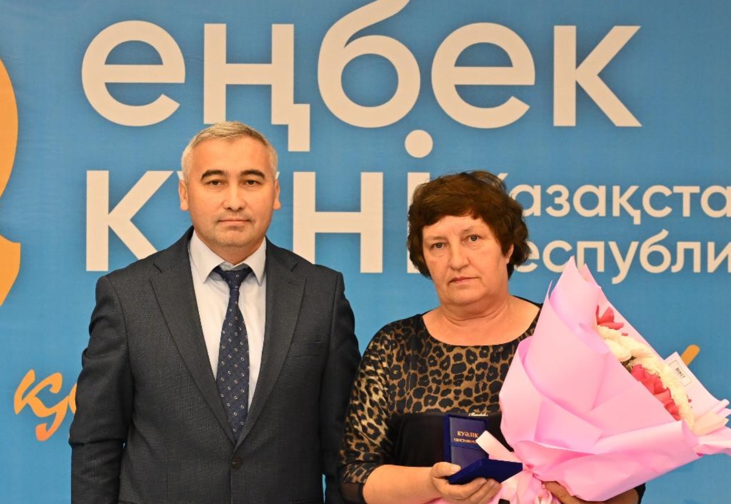 Көк түсті баннер алдында тұрған, қолына гүл шоғы мен куәлік ұстаған әйел мен костюм киген ер адам.