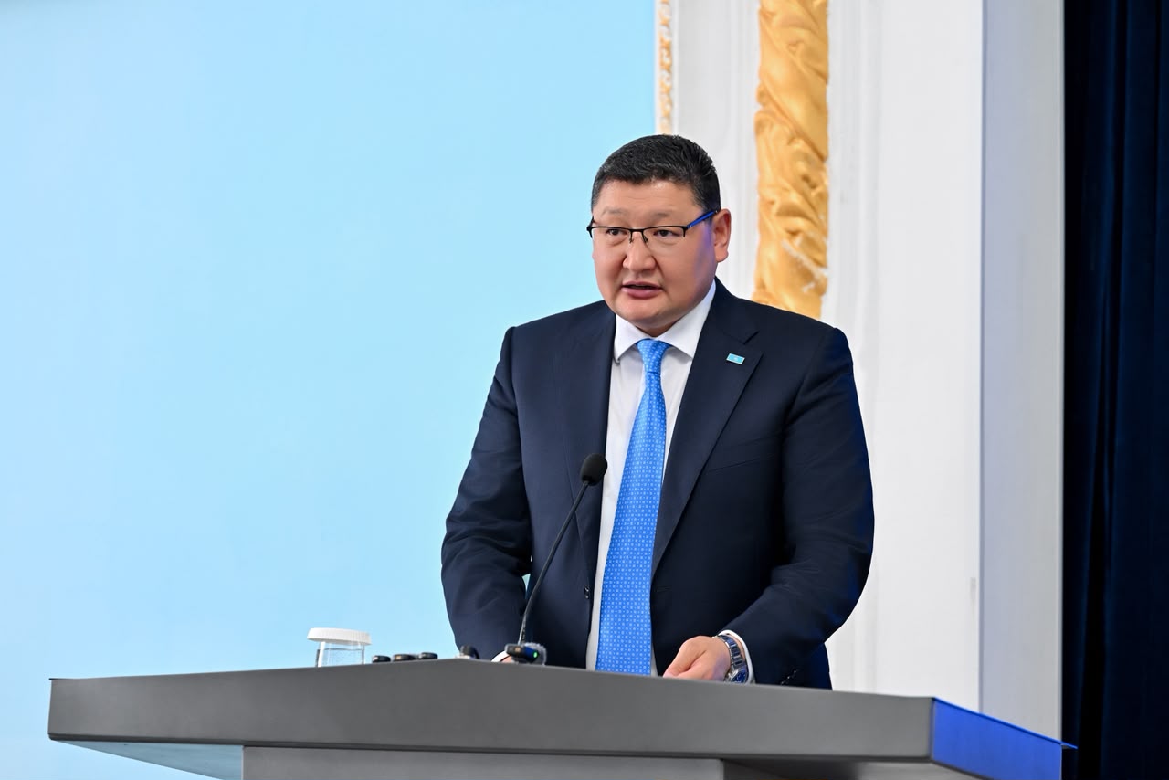 Көзілдірік пен көк галстук киген, трибунада сөйлеп тұрған ер адам.