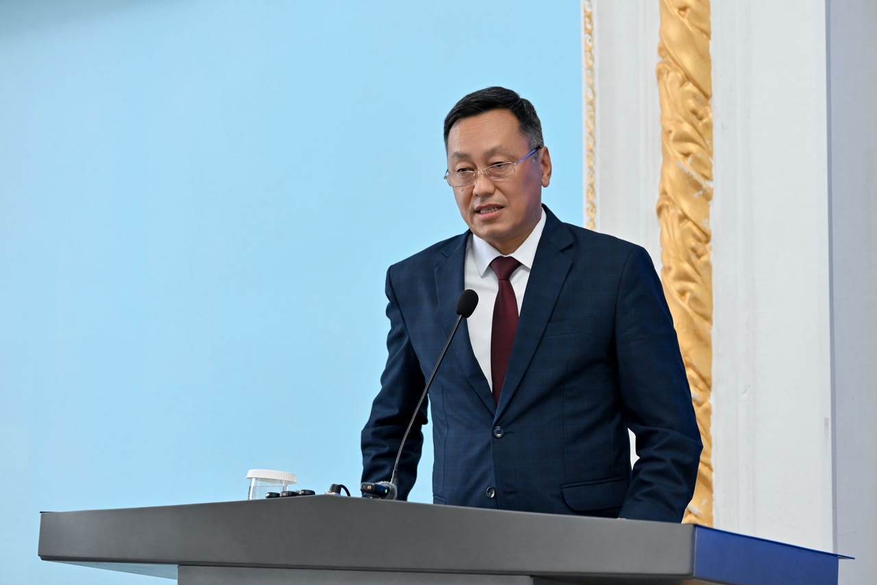 Көзілдірік пен костюм киген ер адам трибунада сөйлеп тұр.