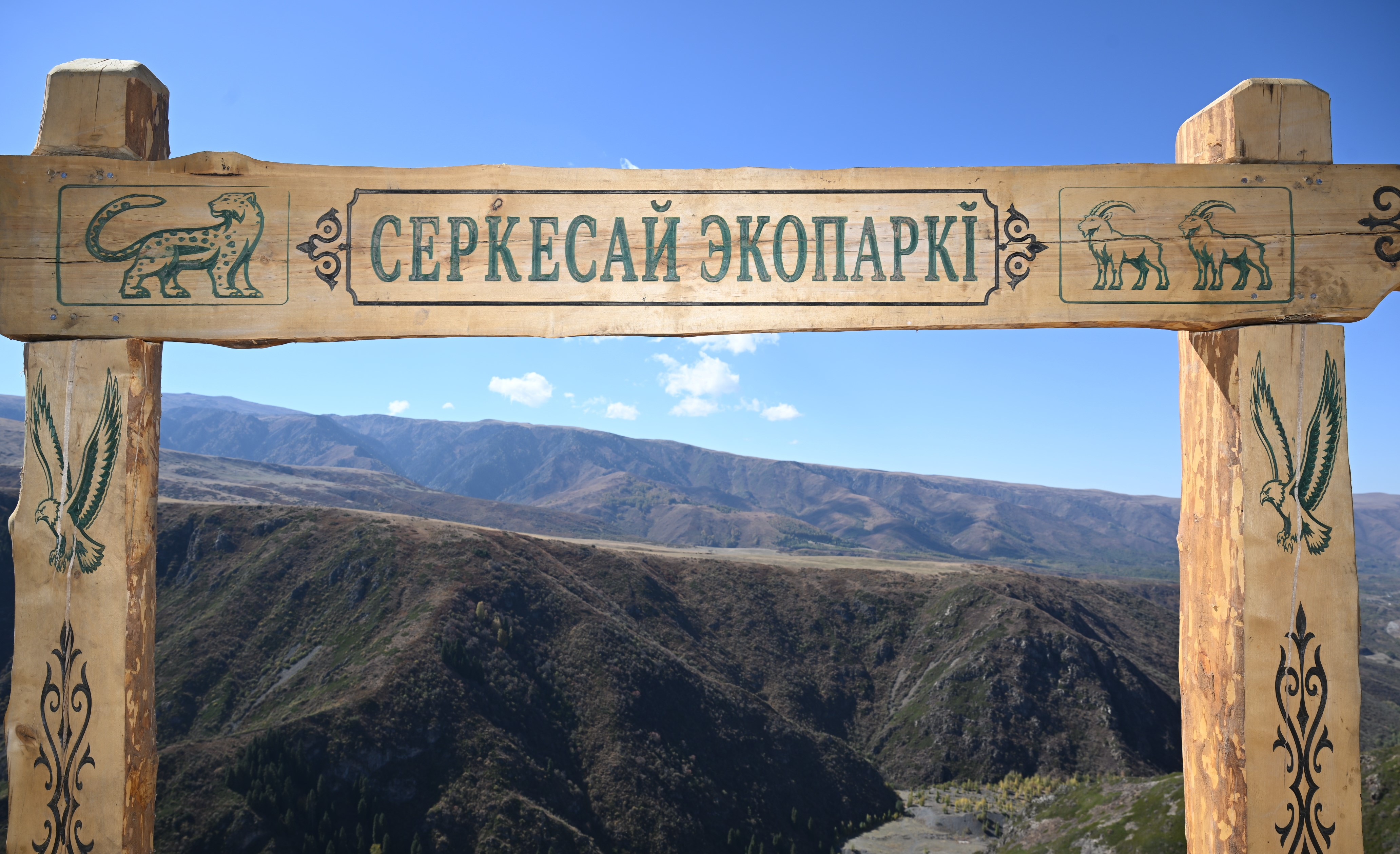 Деревянная арка с надписью «СЕРКЕСАЙ ЭКОПАРКІ» и резьбой в виде животных на фоне горных пейзажей.