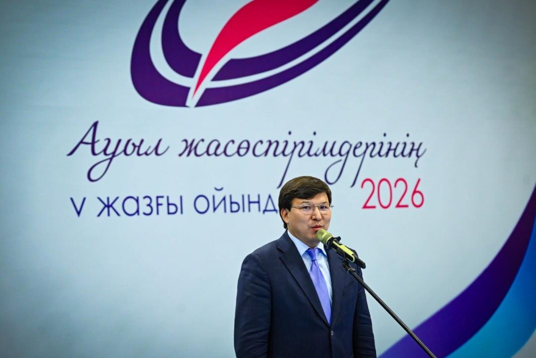 «Ауыл жастарының V жазғы ойындары-2026» баннерінің алдында сөйлеп тұрған костюм киген ер адам.